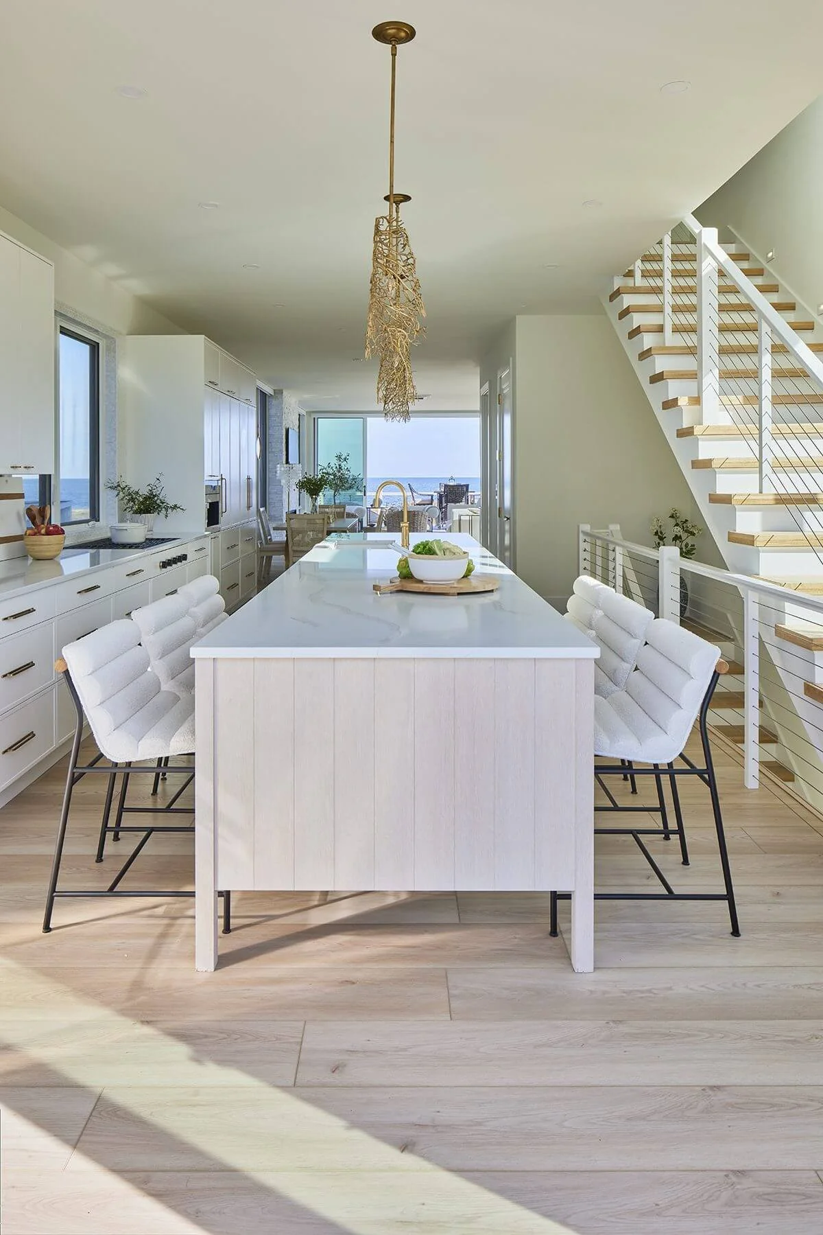 Modern, bright kitchen and dining area with a large white island, white chairs, multiple windows, and a staircase with wooden steps and white railings, overlooking an outdoor view.