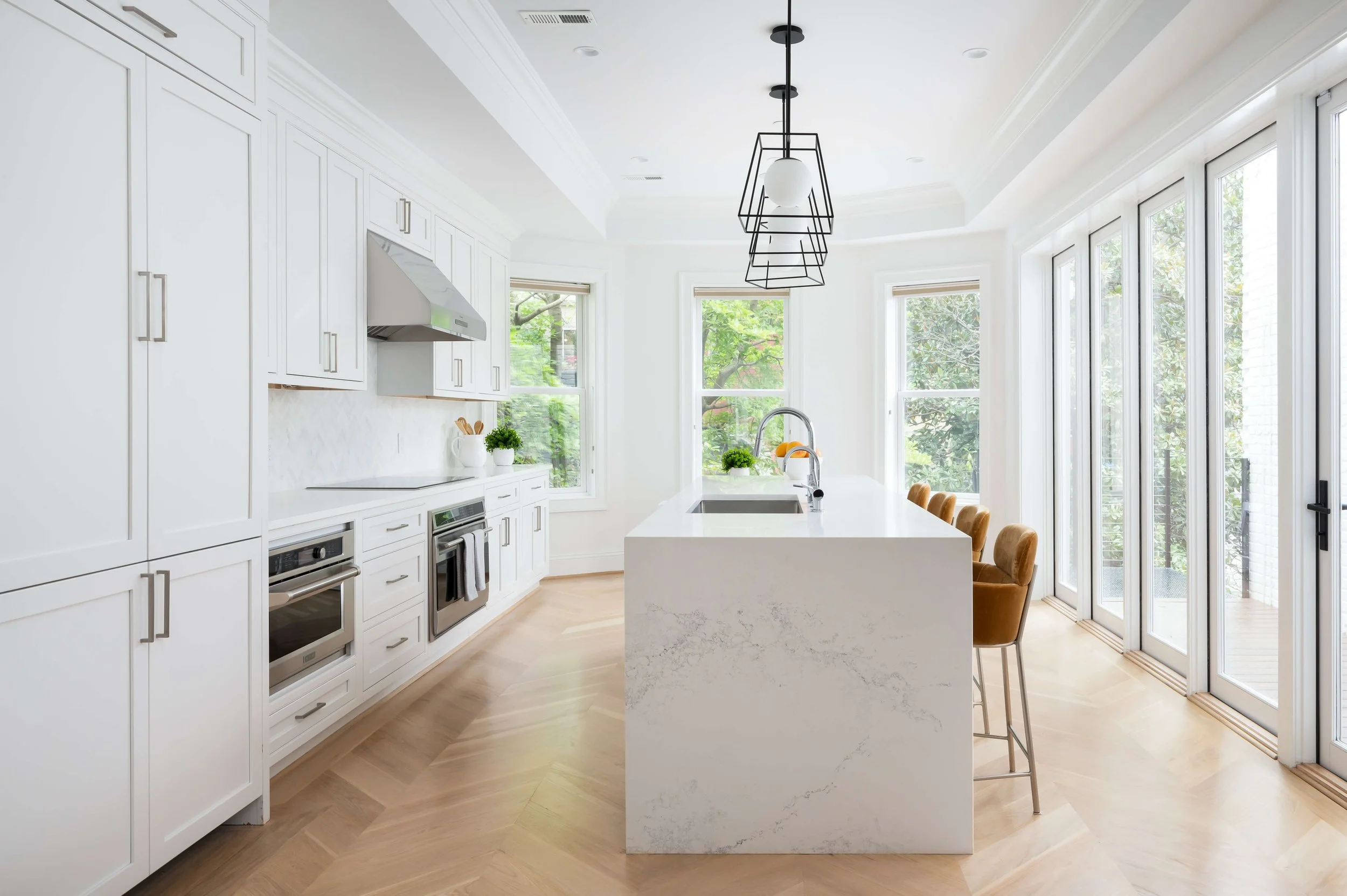 Modern white kitchen with large island, stainless steel appliances, and floor-to-ceiling windows with greenery outside.