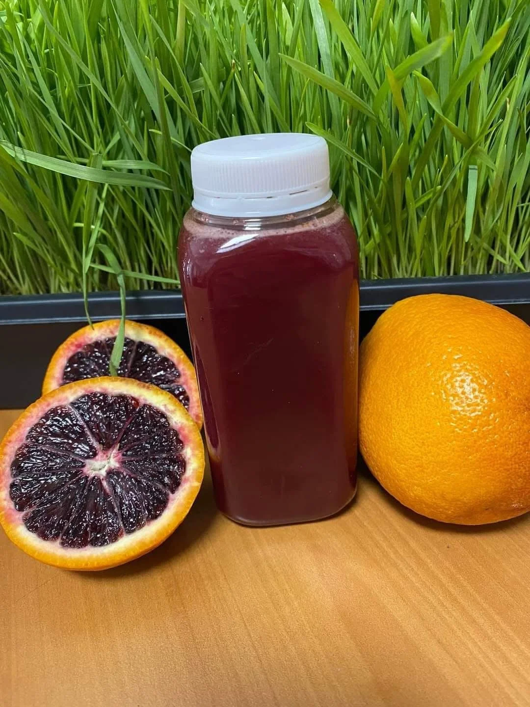 A juicer bottle filled with dark red juice, placed on a wooden surface next to halved blood oranges and a whole orange, with green grass in the background.