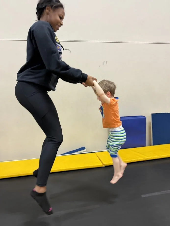A woman and a young boy playing on a trampoline, holding hands and jumping together indoors.