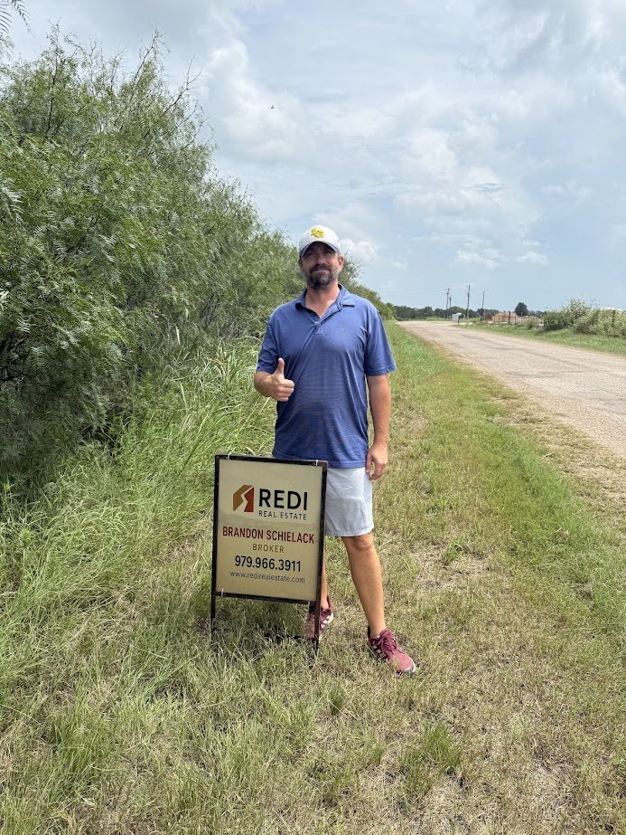 Man standing next to a sign for Redwood Real Estate on a grassy roadside, giving a thumbs-up and wearing a blue polo shirt, shorts, and a white cap.