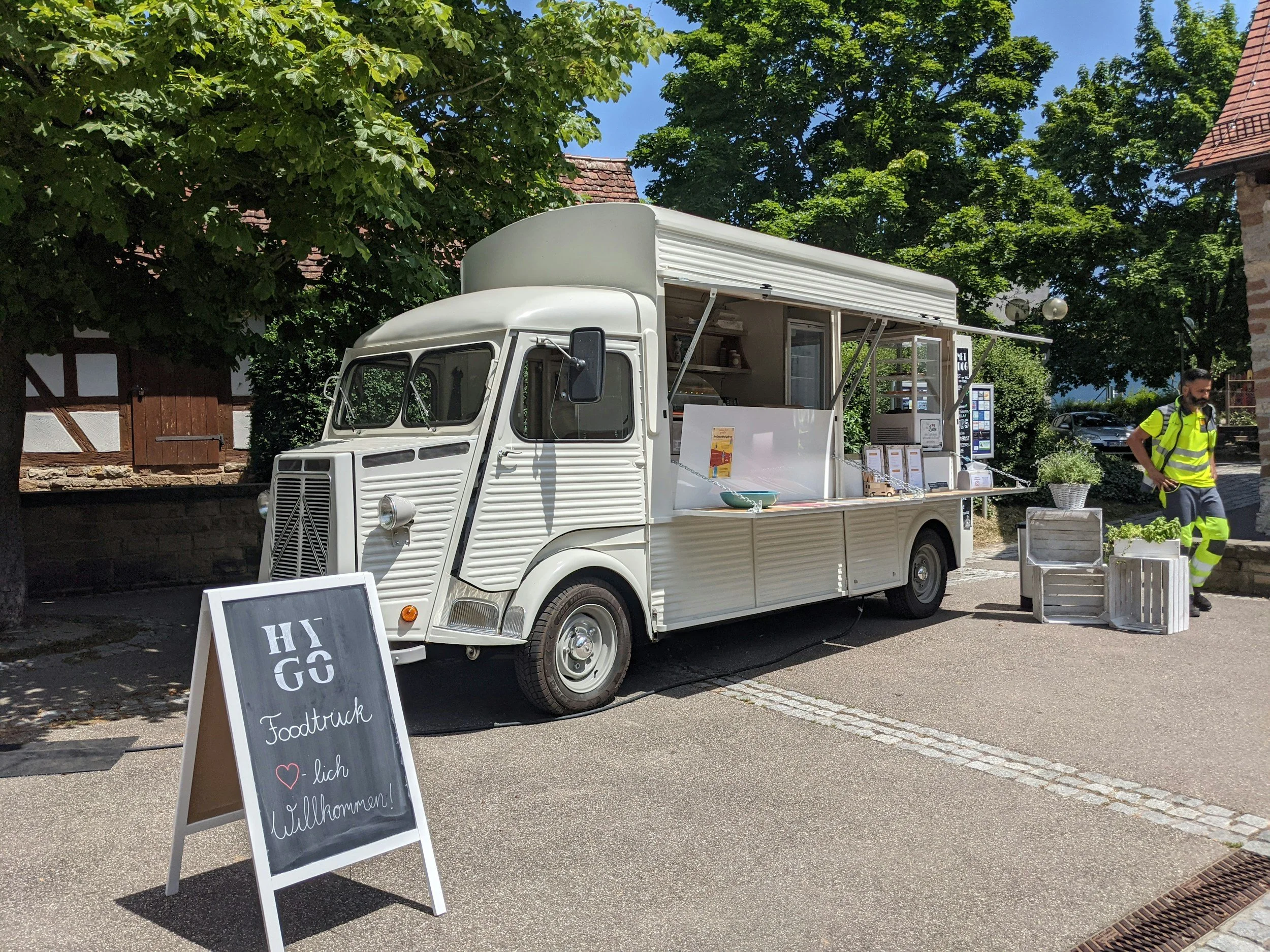 A vintage white food truck parked on a street near trees and houses. The truck has a serving window with menus and a menu board nearby. An A-frame sign in front reads 'HI GO Foodtruck ♥ Dich Willkommen!'. A man in a reflective yellow vest stands nearby.