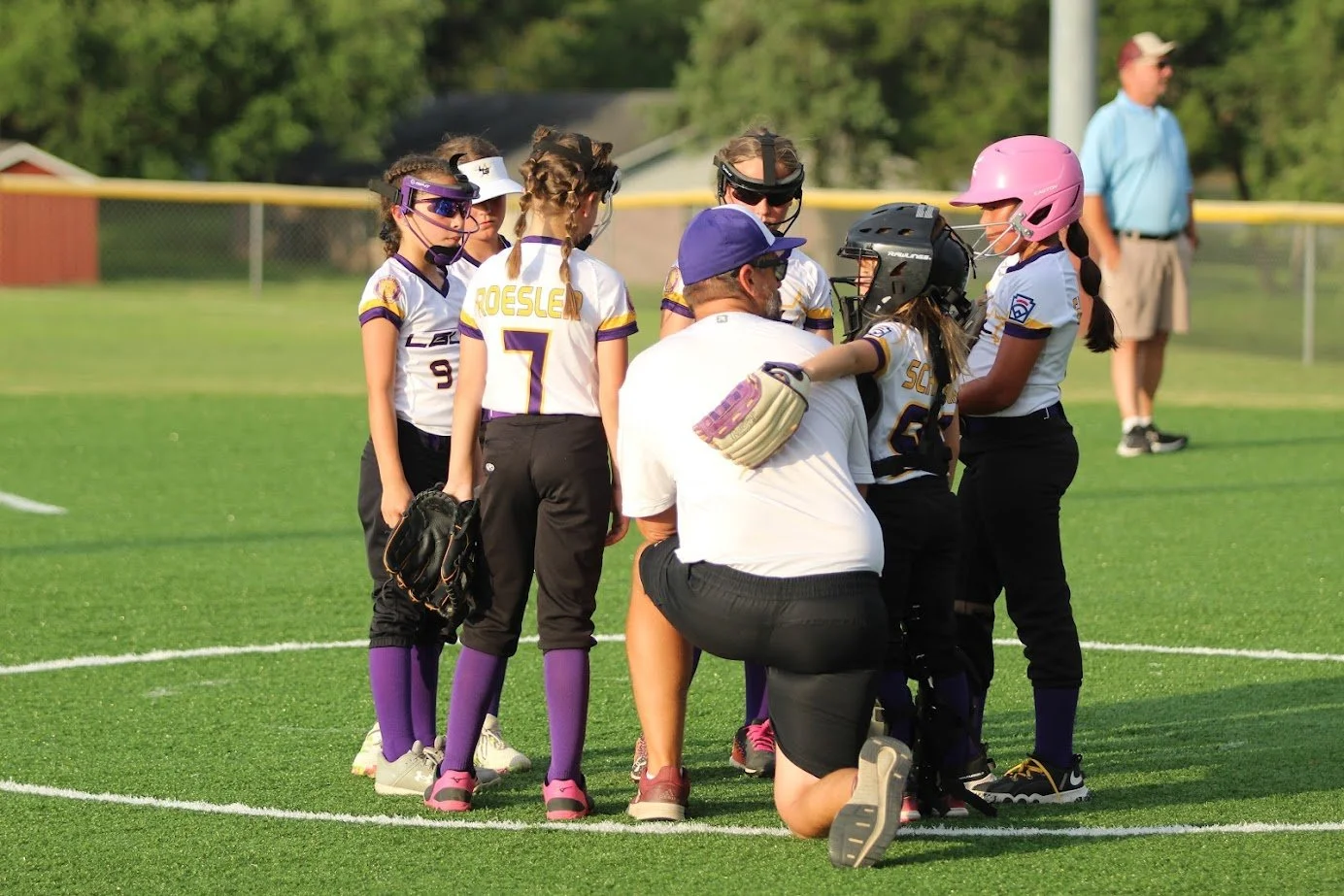 Softball team huddled around coach on field during game, wearing purple, white, black uniforms and protective gear.