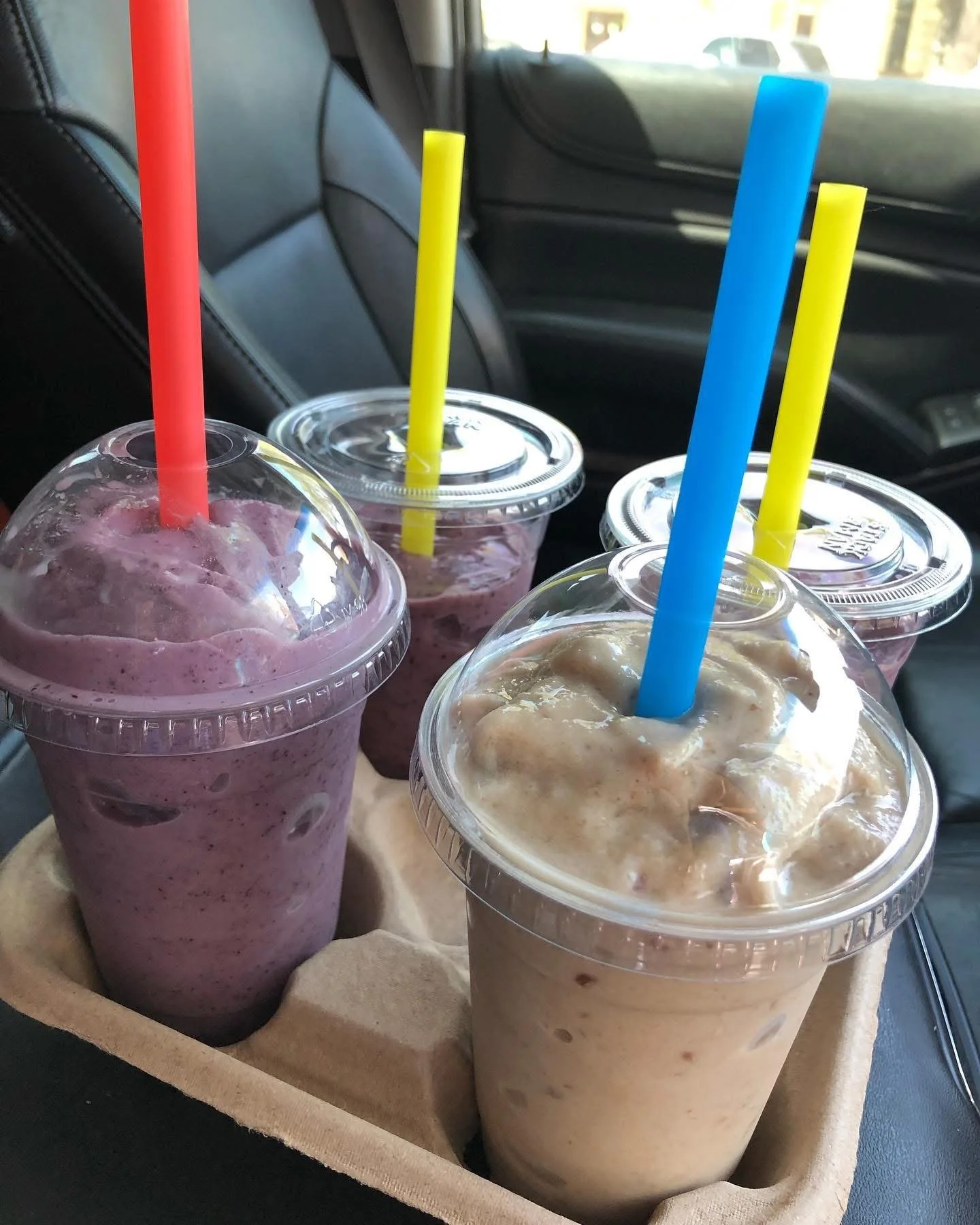 Four large iced beverages in plastic cups with colorful straws, placed in a cardboard drink holder inside a vehicle.