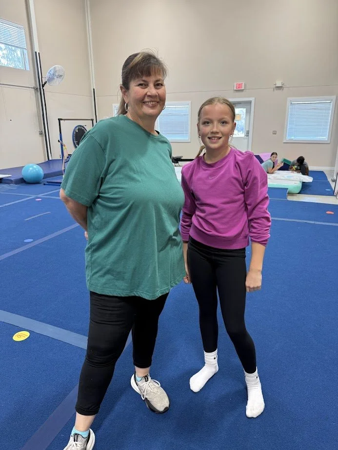 Two females, a woman and a girl, standing on a blue gym floor inside a gym or training facility, smiling at the camera.