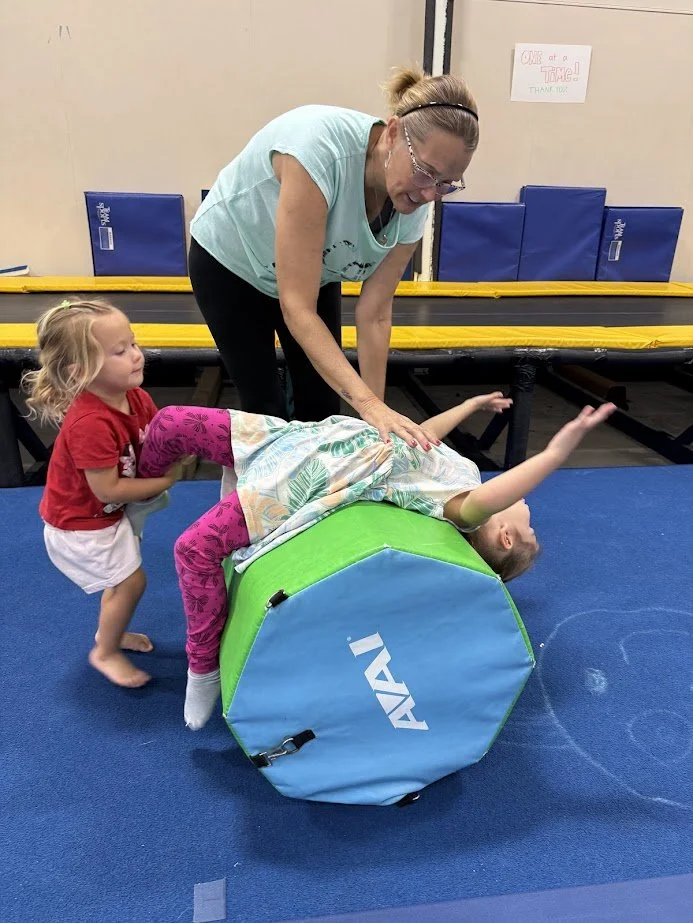 A woman and two young girls playing on a padded gymnastics mat at an indoor trampoline park. One girl is lying on a green and blue foam roller while the other girl helps her balance, and the woman is assisting and smiling during their activity.