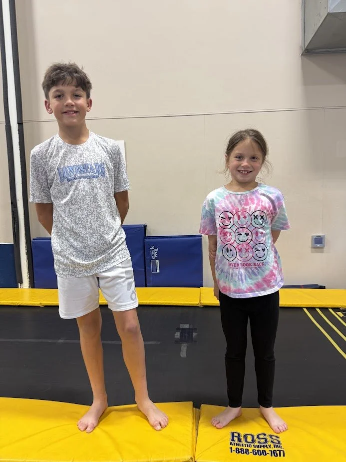 Two children, a boy and a girl, are standing barefoot on yellow foam padding inside a gym. The boy is wearing a gray t-shirt and white shorts, smiling, with hands behind his back. The girl is wearing a tie-dye t-shirt with smiley faces and black leggings, also smiling.