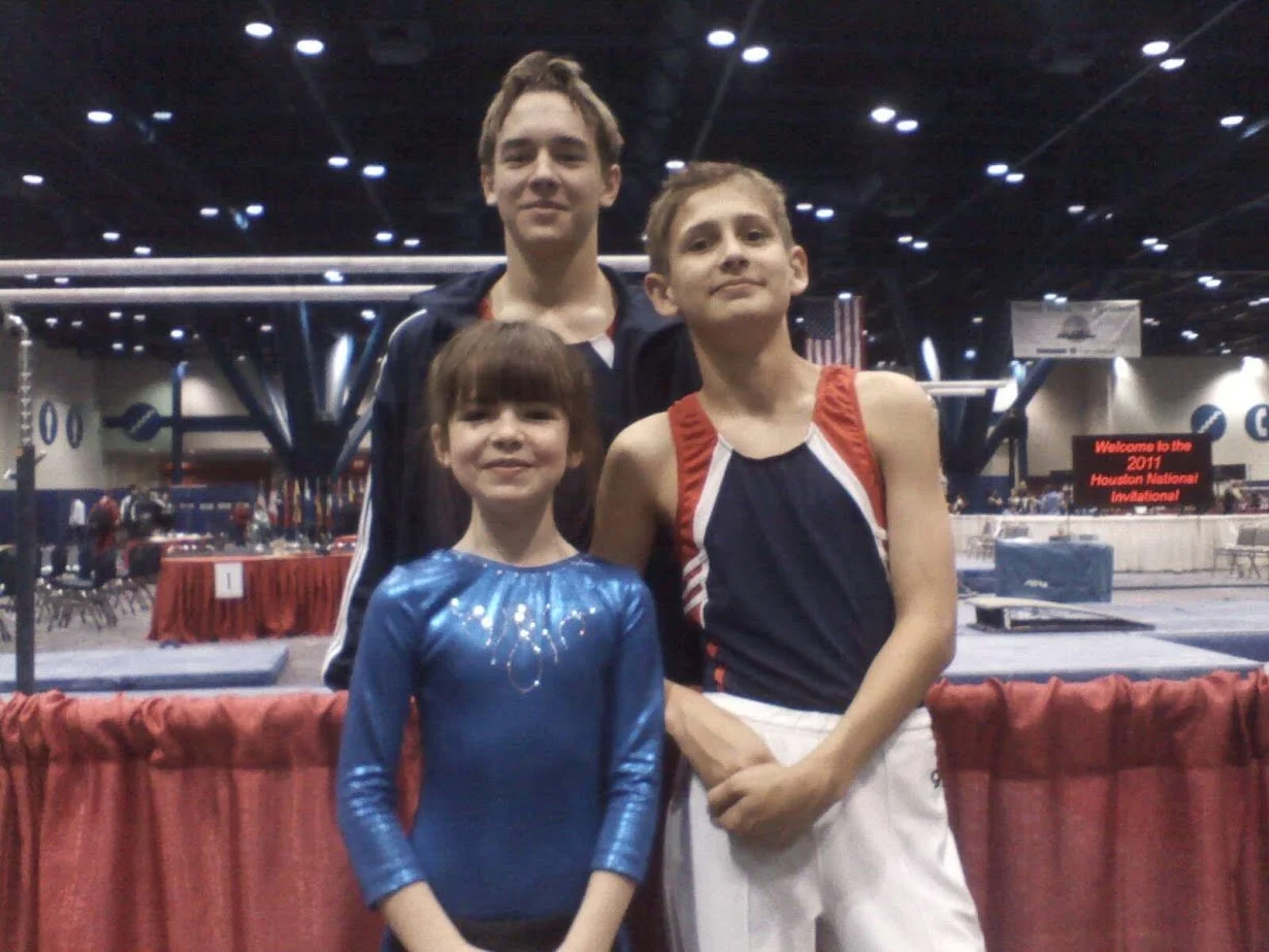 Three children standing in front of a gymnasium at the 2011 Houston National Invitational event, with a red curtain in the foreground and a sign in the background welcoming attendees.
