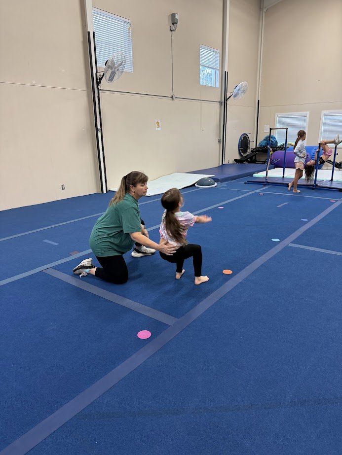 A woman coaching a young girl during a gymnastics class inside a gym. The girl is jumping or running on a blue mat with colorful markers. In the background, two other children are practicing at a low horizontal bar.