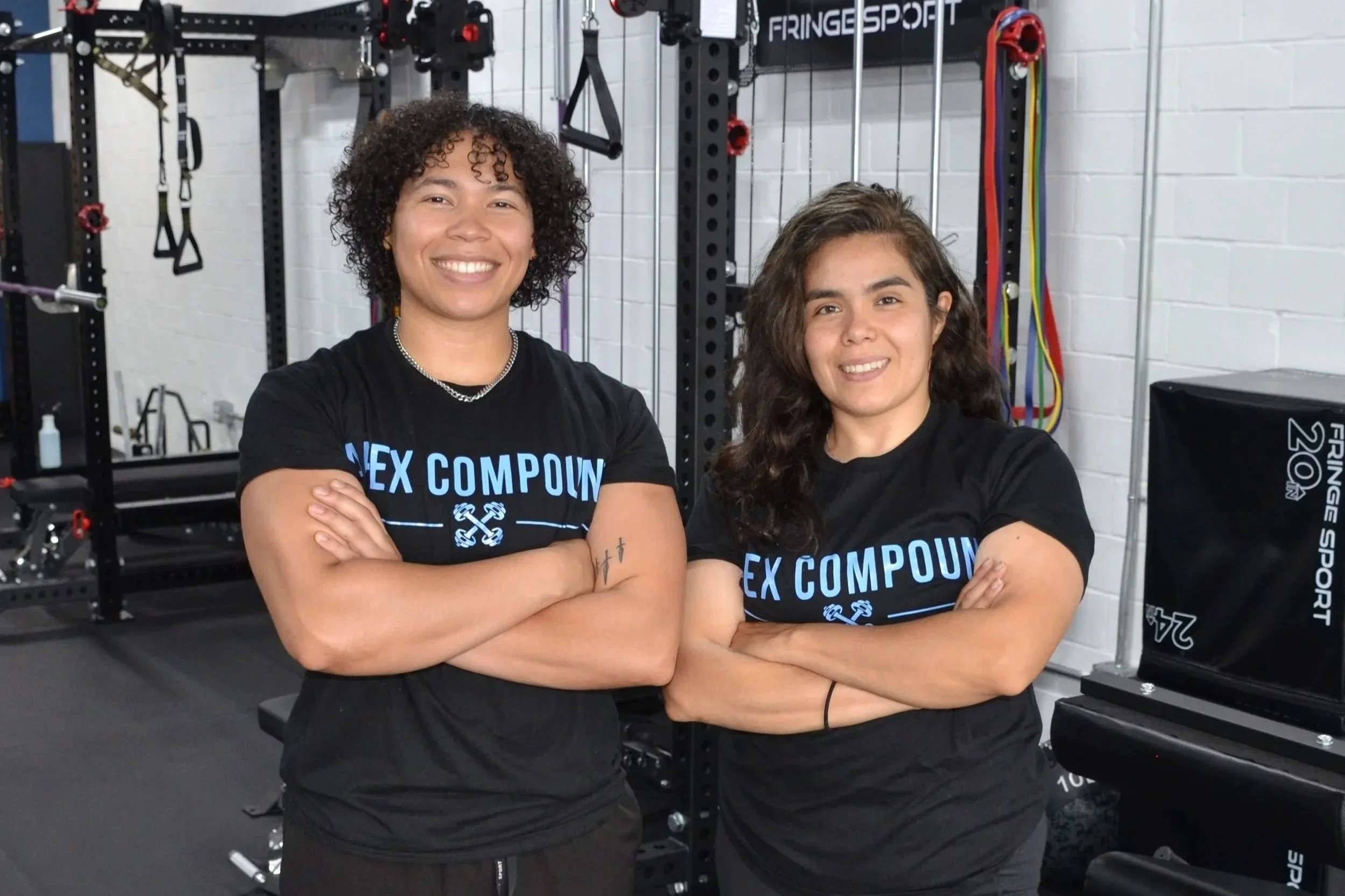 Two women with crossed arms smiling at gym, wearing black T-shirts with blue text, fitness equipment in background.