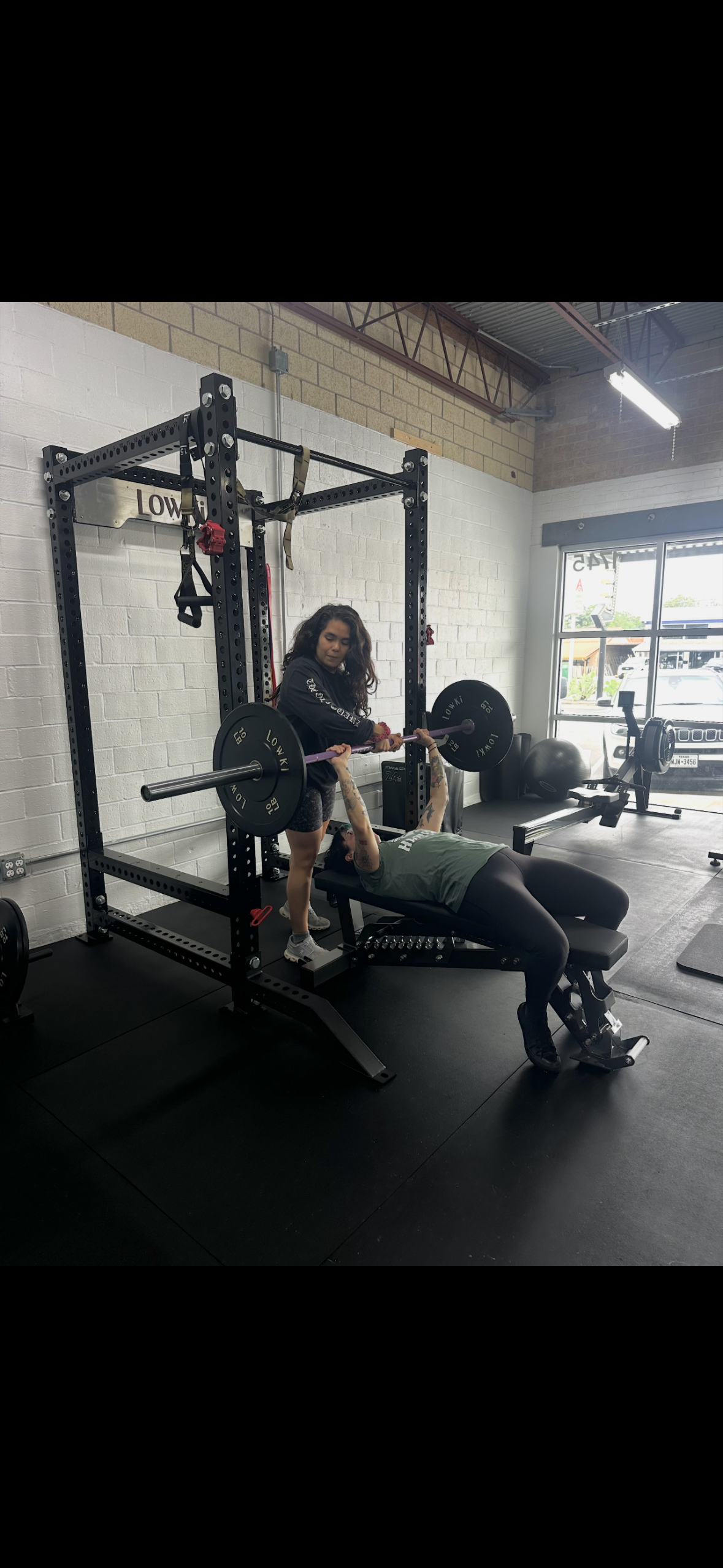 A person lifting weights while another person, possibly a trainer, provides assistance or coaching in a gym.