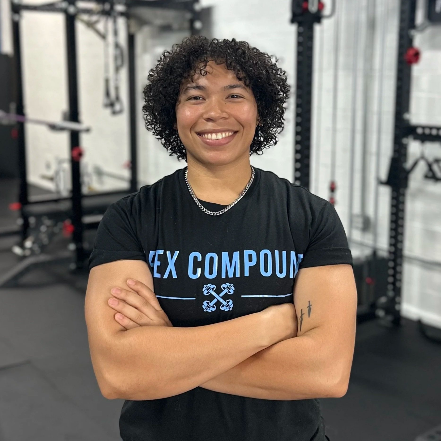 A person with curly hair smiling with arms crossed in a gym setting, wearing a black shirt and a silver chain, surrounded by gym equipment.