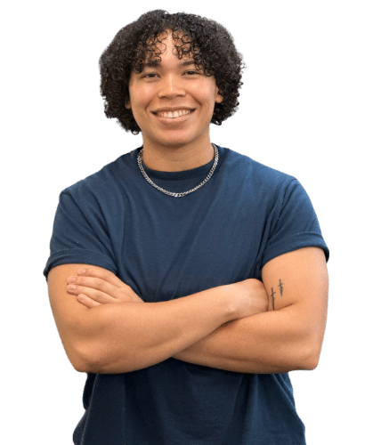 A young person with curly hair smiling, wearing a blue t-shirt, a silver chain necklace, and an arm tattoo, crossing their arms.