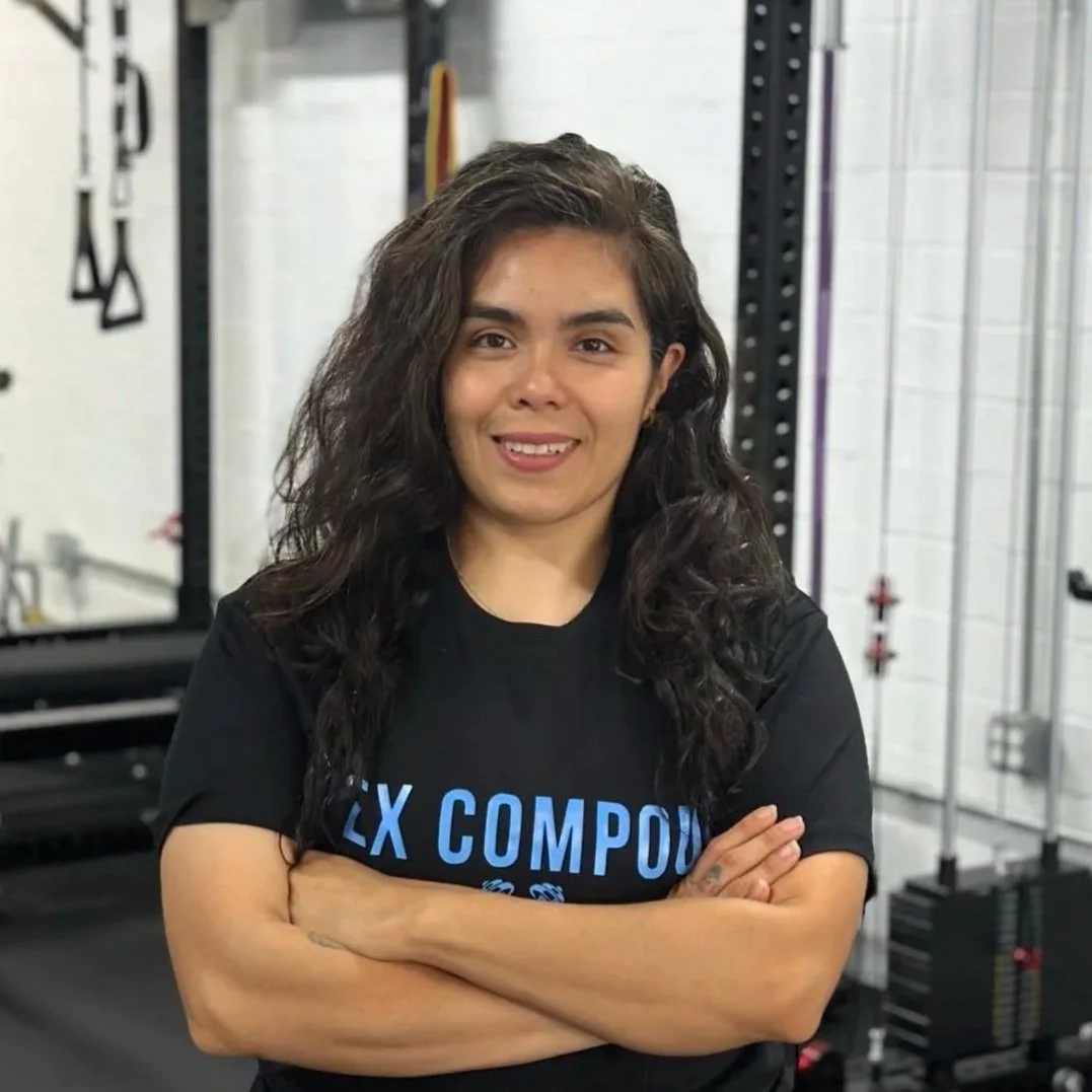 A young woman with curly dark hair standing with arms crossed in a gym, wearing a black t-shirt with blue text.