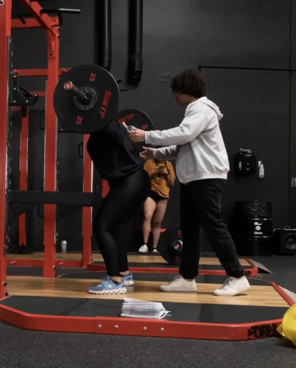 A person lifting weights while another person, possibly a trainer, provides assistance or coaching in a gym.