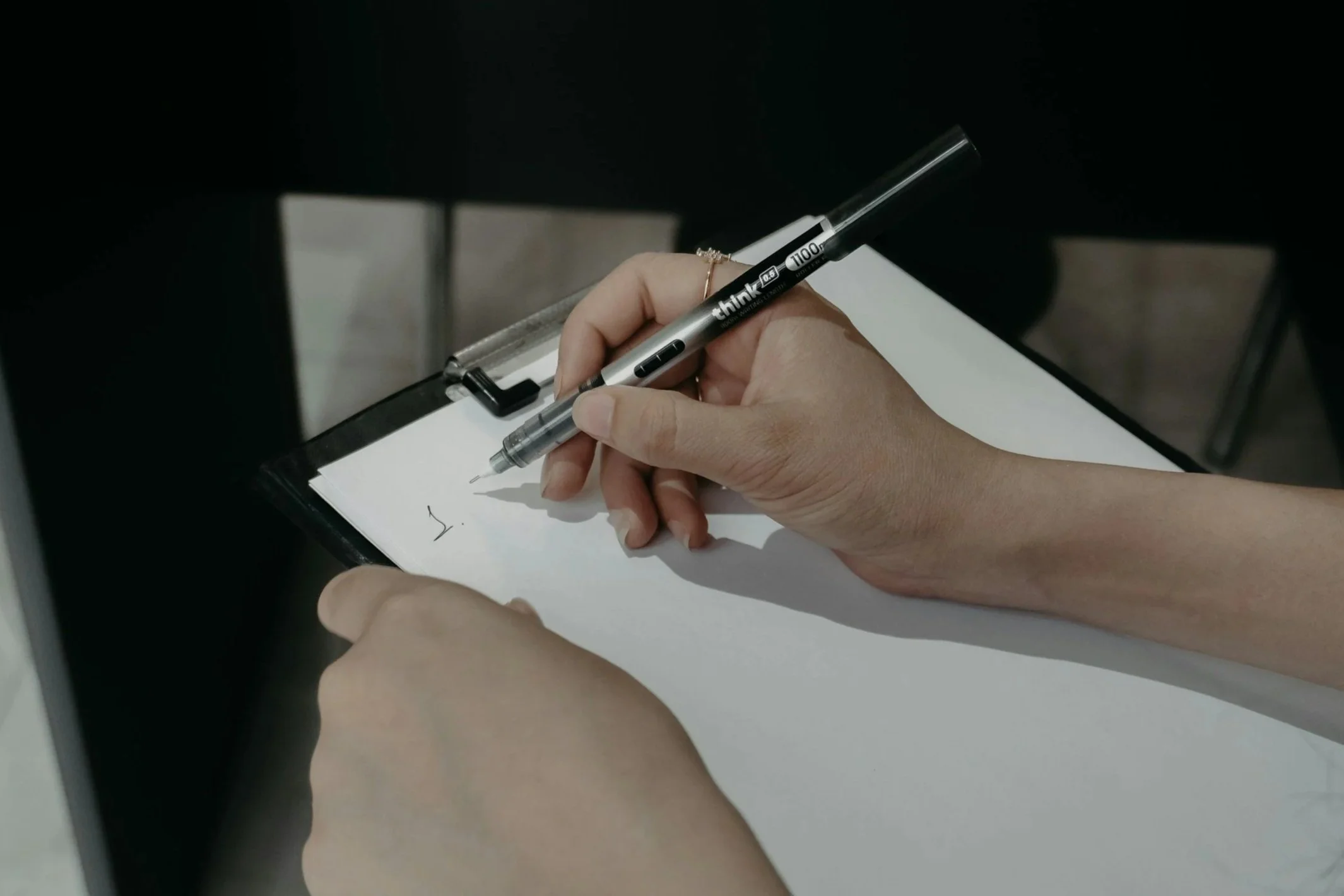 Person writing on a clipboard with a black and gray pen in a well-lit environment.