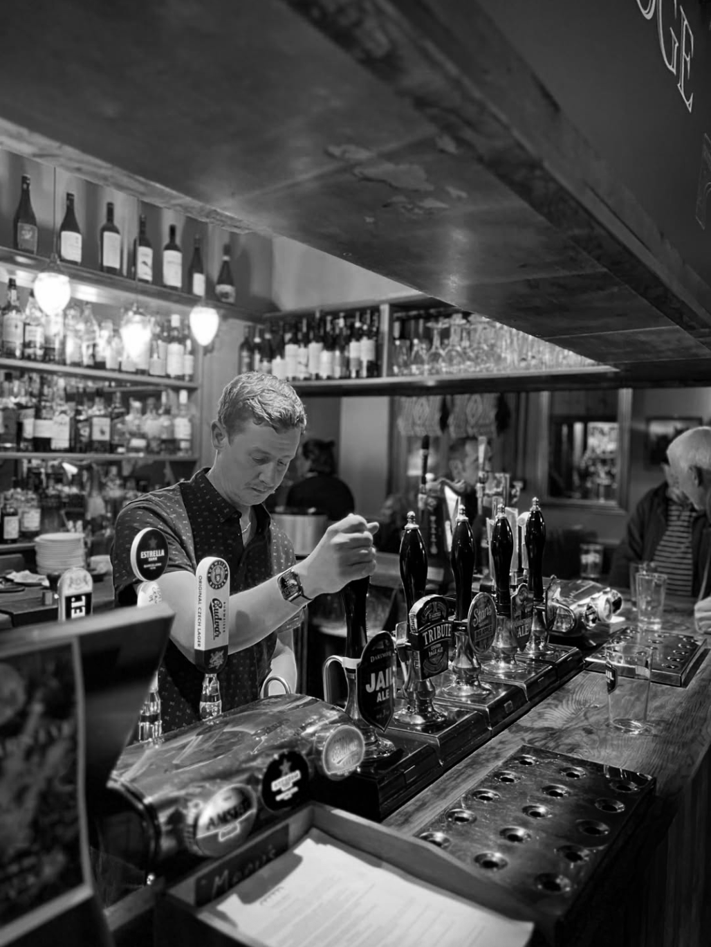 A man working behind a bar with multiple beer taps in front of him, shelves of bottles behind, and customers seated in the background.