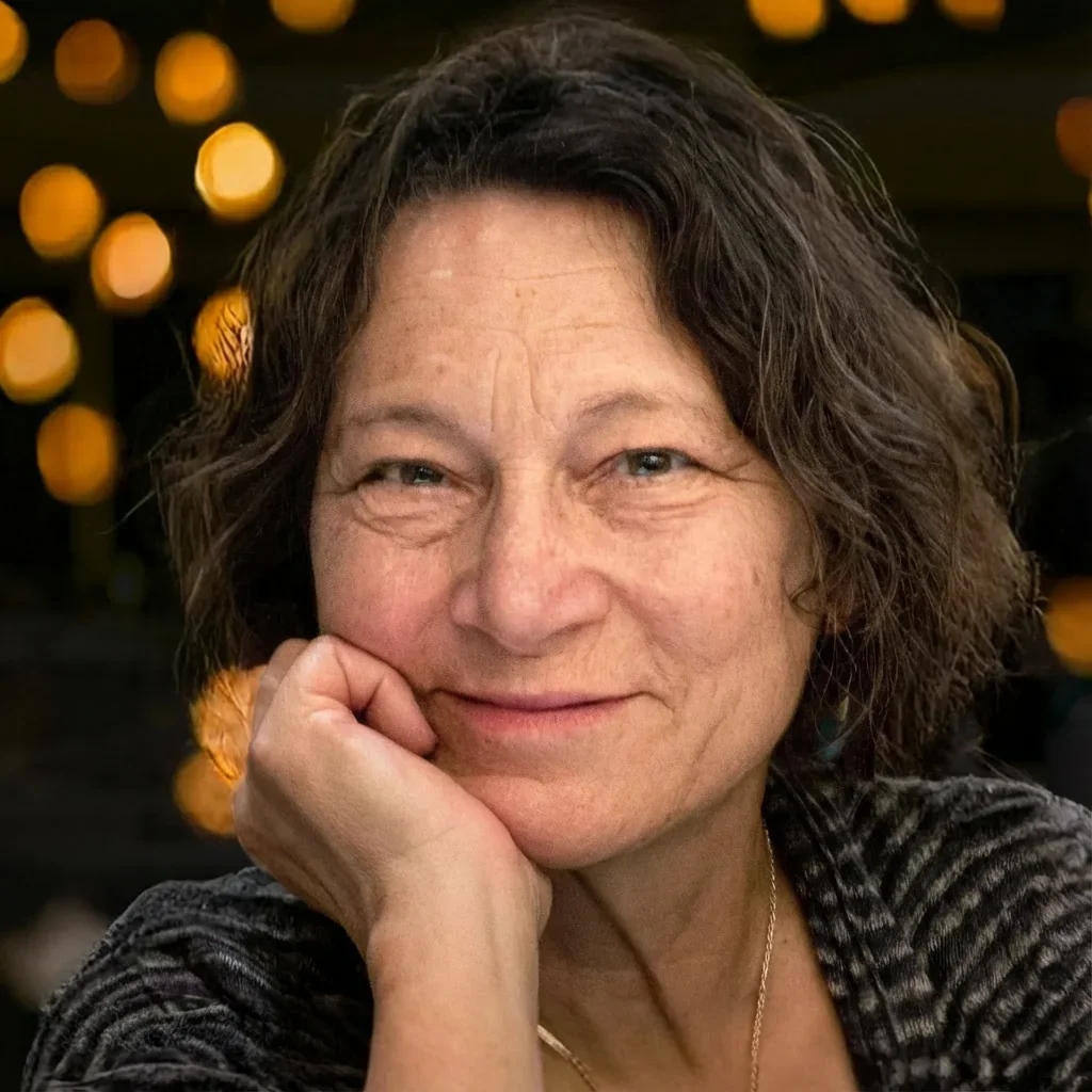 Close-up of a middle-aged woman with curly brown hair, smiling gently, resting her chin on her hand, in an indoor setting with yellow decorations in the background.