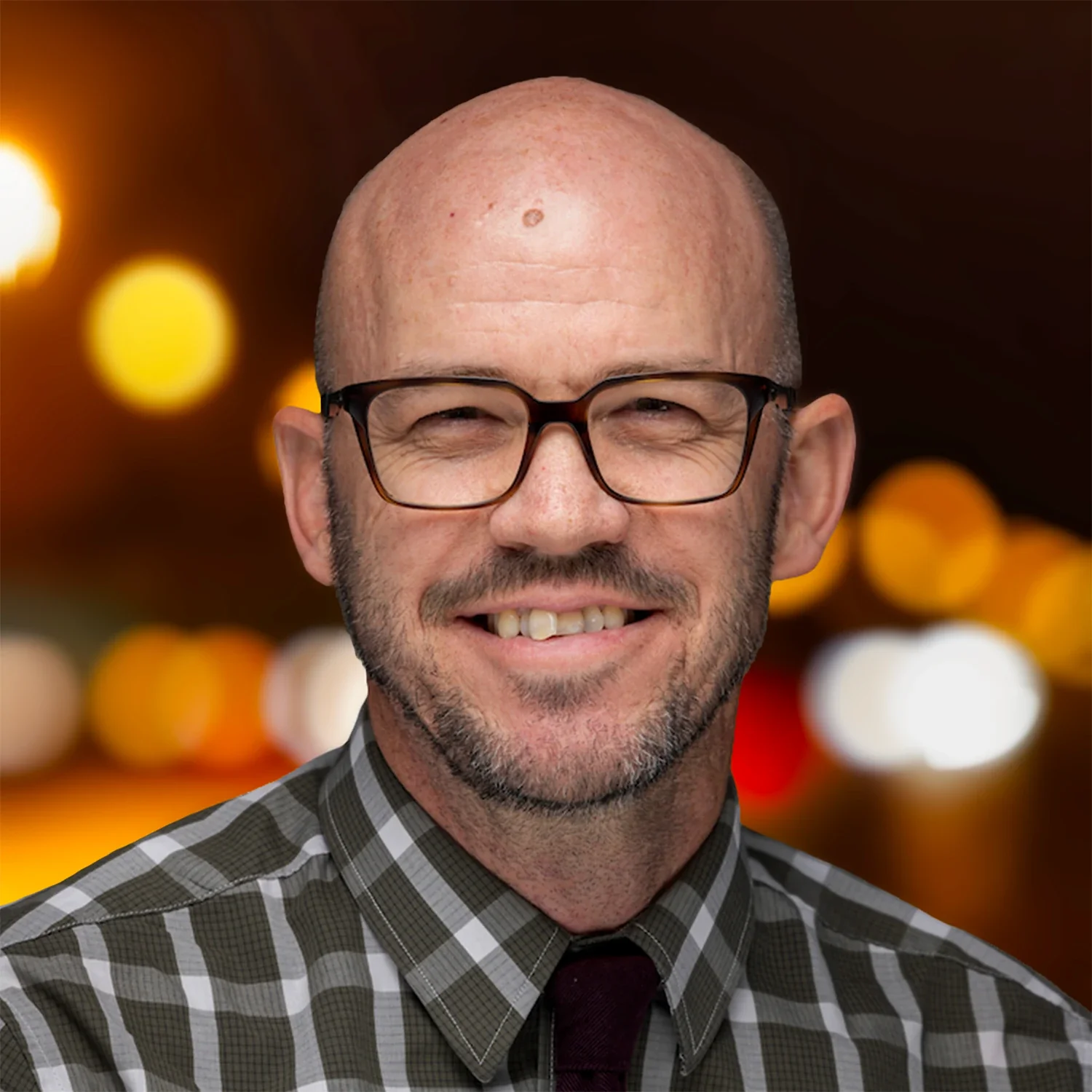 Portrait of Brock Jones smiling, wearing glasses, a plaid button-down shirt, and a dark tie against a softly blurred background of warm lights.
