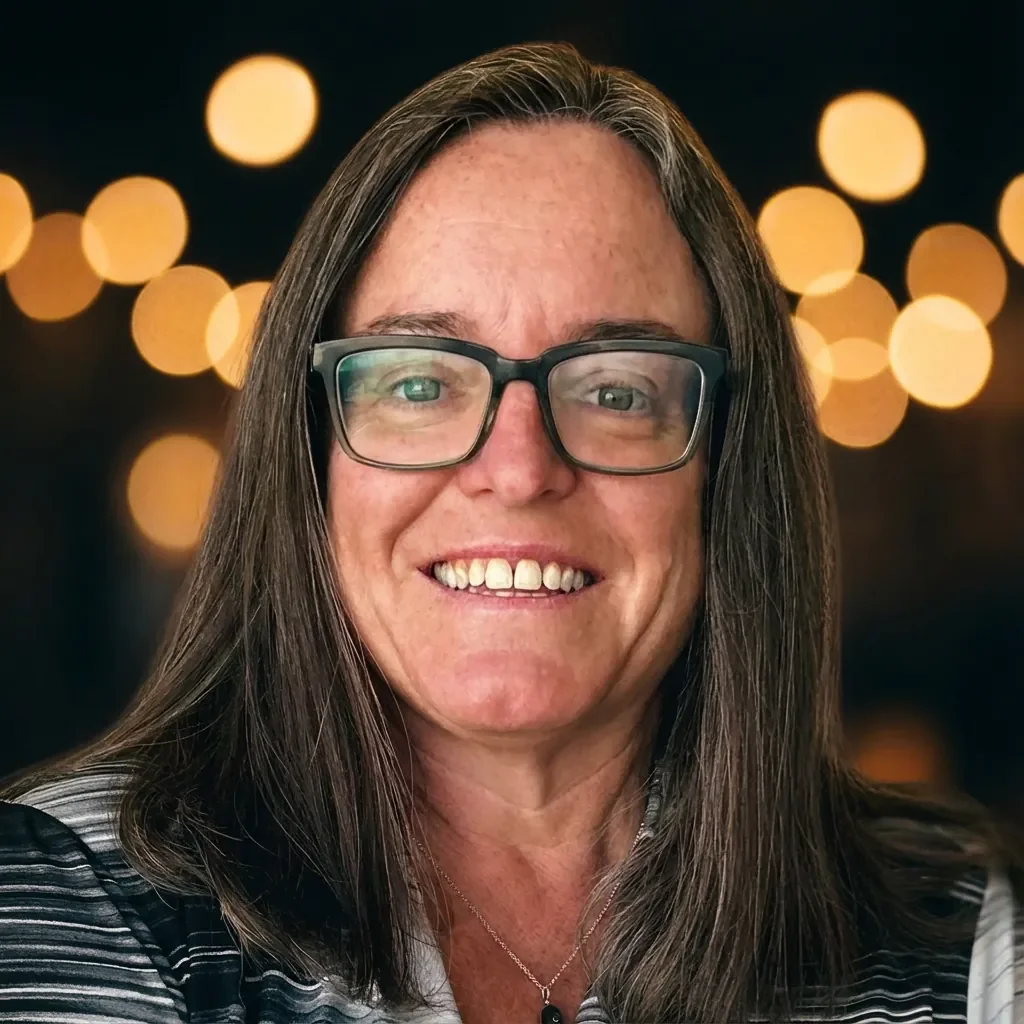 A woman with long brown hair, glasses, and a light-colored shirt, smiling against a blurred background of city lights at night.