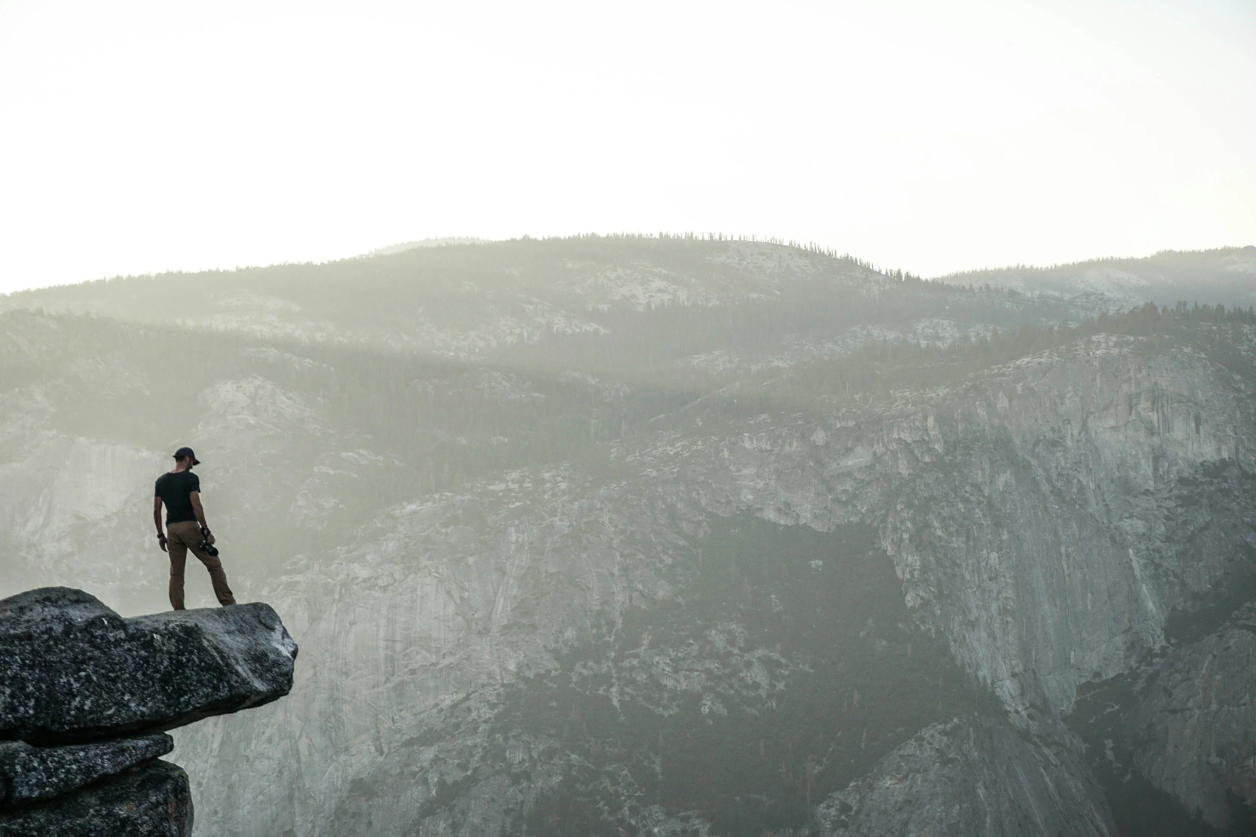 A person standing on the edge of a rocky ledge overlooking a mountainous landscape with steep cliffs and pine trees, during daylight.
