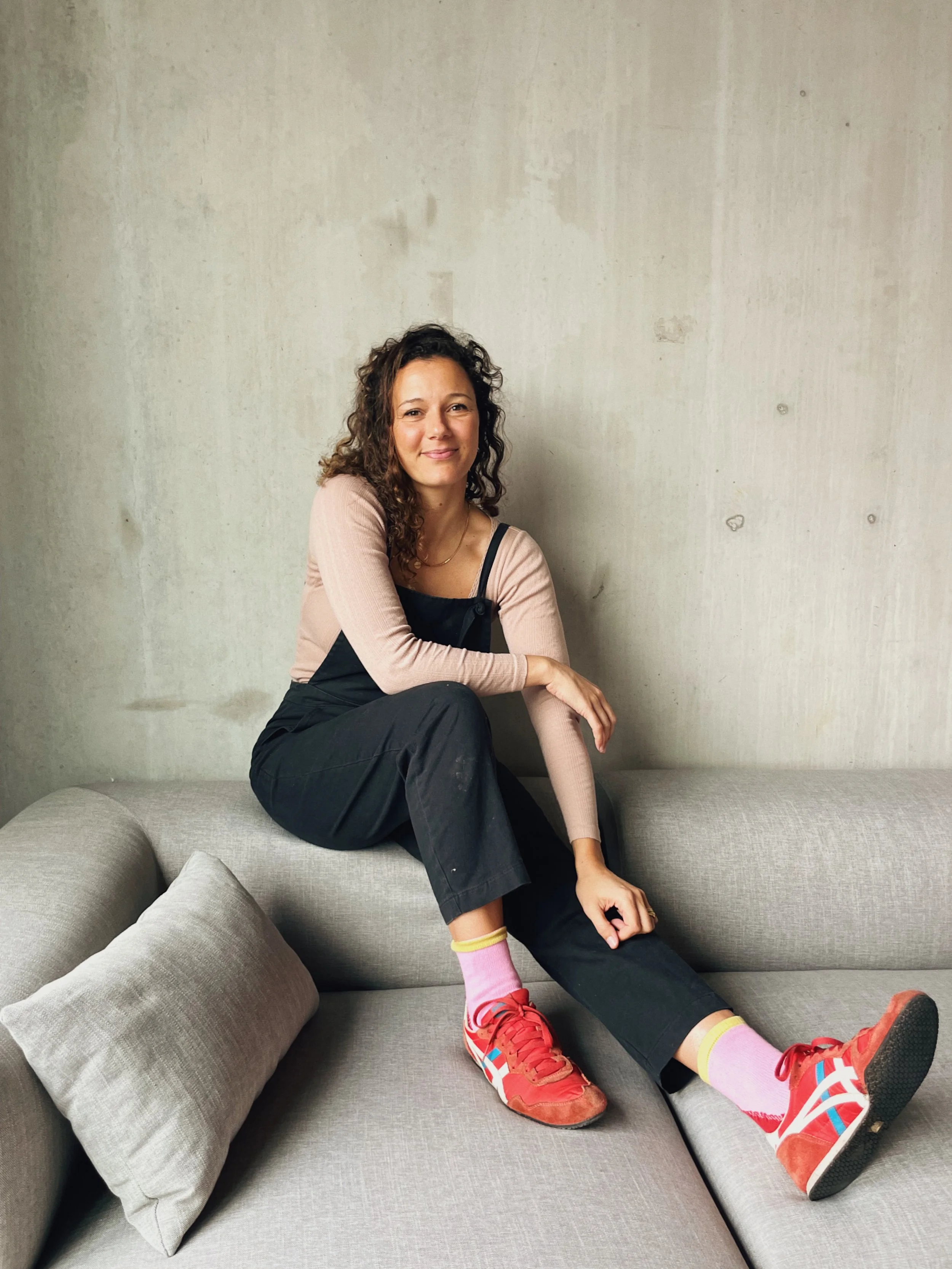 A woman with curly hair sitting on a gray sofa with a plain light-colored wall behind her, wearing pink socks, red sneakers, black overalls, and a beige long-sleeve top, smiling at the camera.