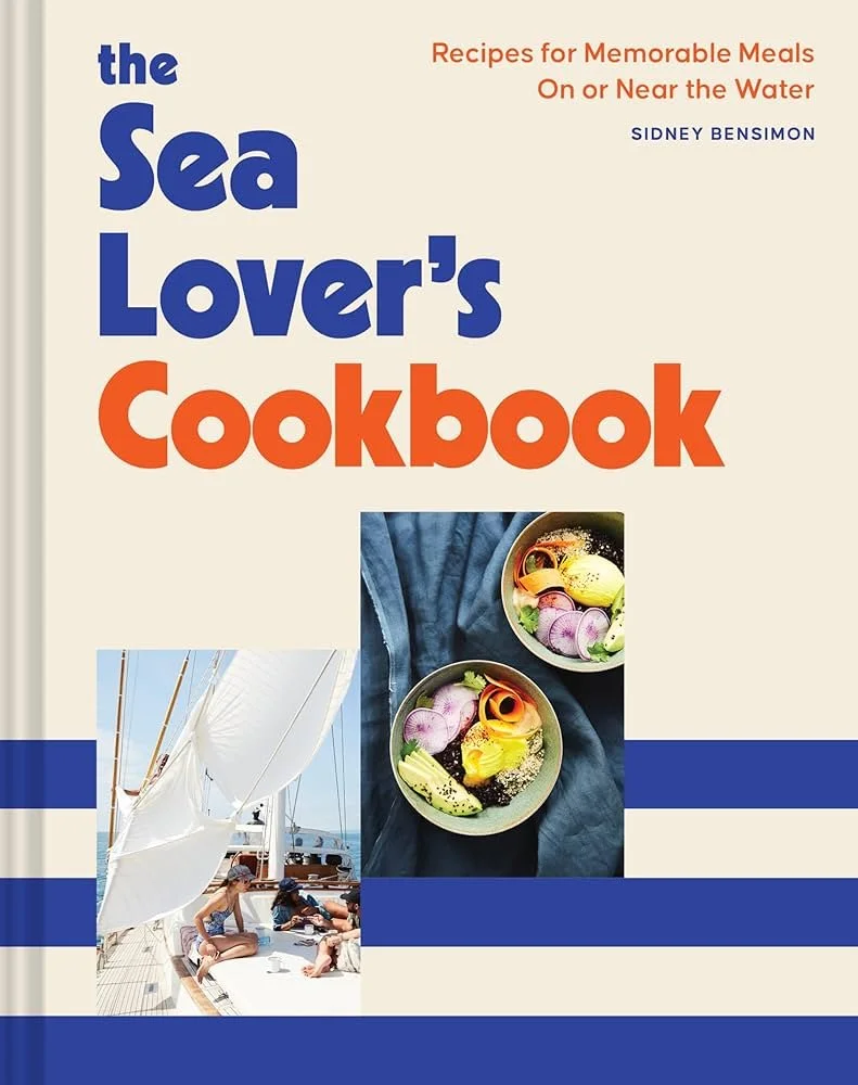 Cover of "The Sea Lover's Cookbook" featuring a sailboat with children playing on deck, and two bowls of salad with vegetables and flowers, set on a blue cloth.