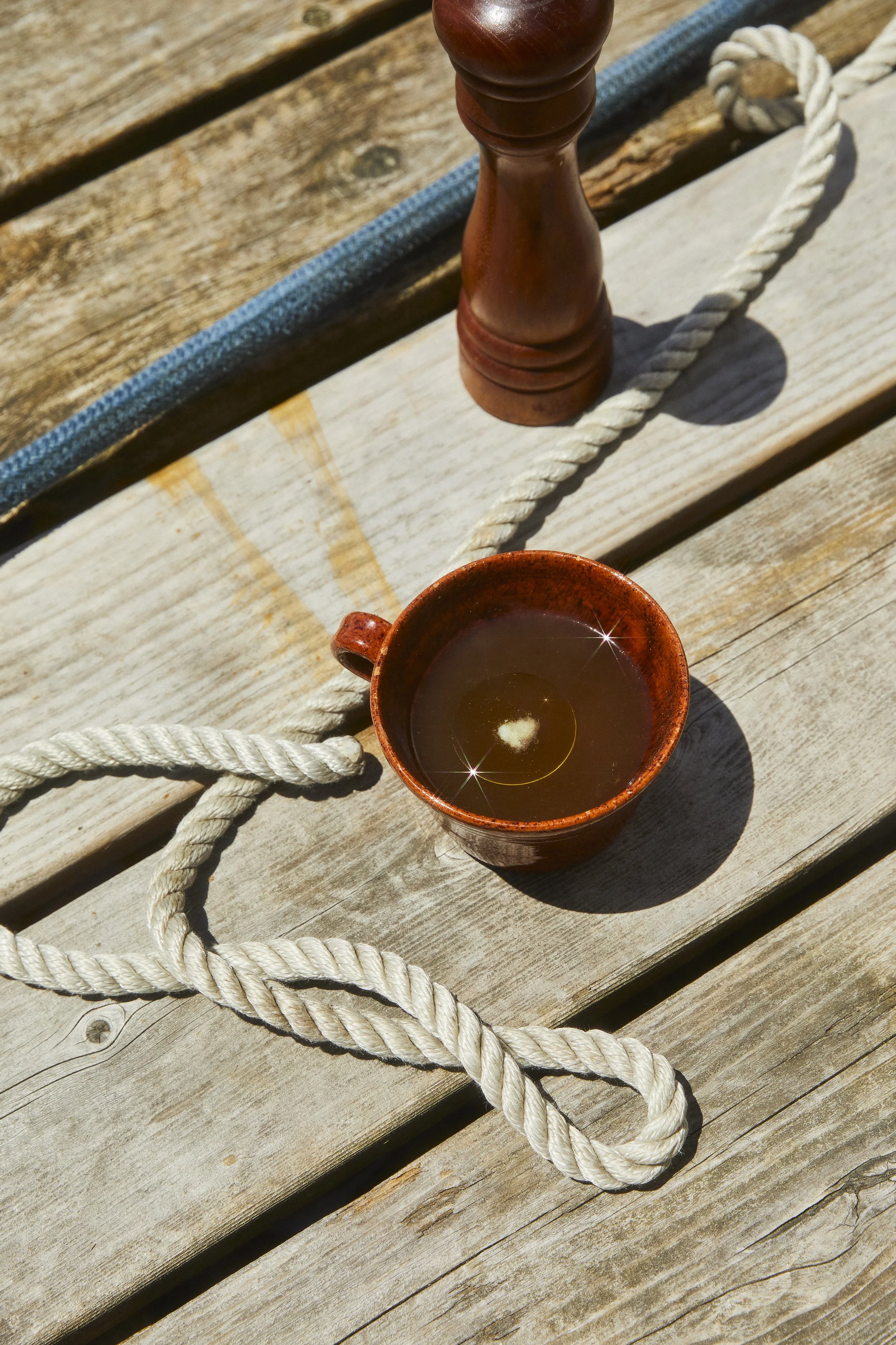 A small, brown ceramic cup filled with tea, with a wood and metal object and a white rope on a weathered wooden surface outdoors.