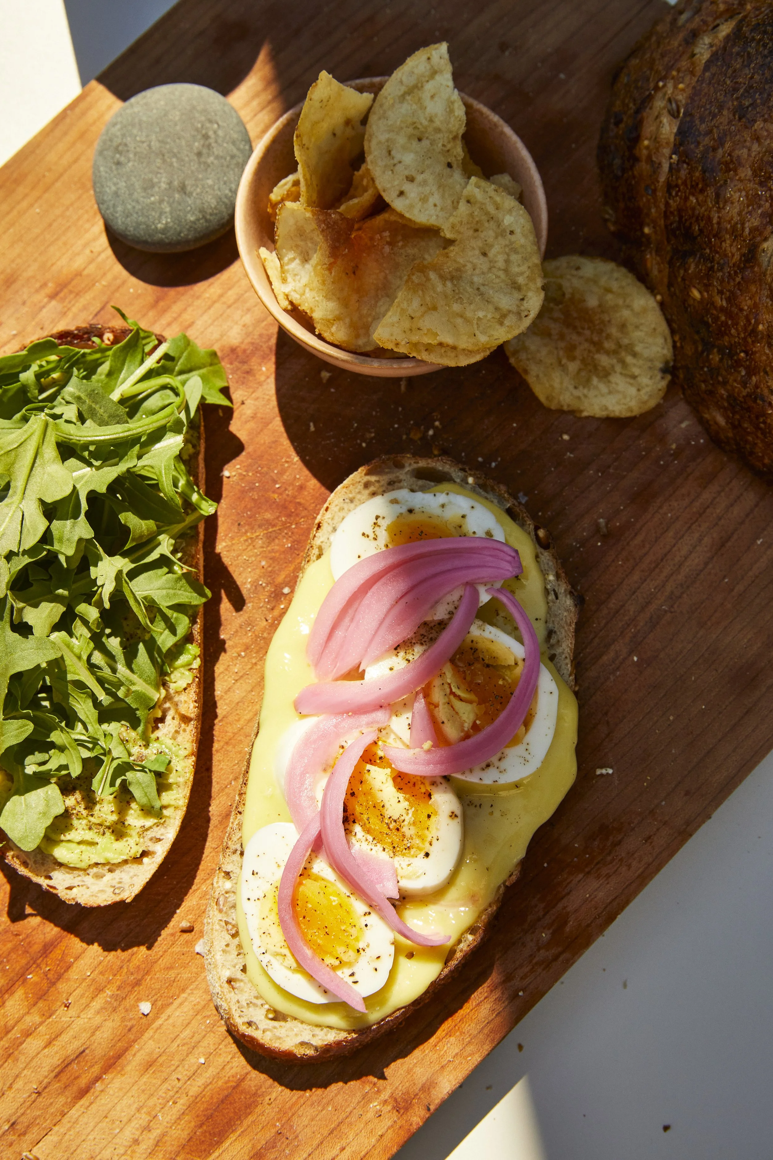 An open-faced sandwich with sliced boiled eggs, melted cheese, pickled red onions, and black pepper on toasted bread, with a side of fresh arugula and potato chips on a wooden cutting board.