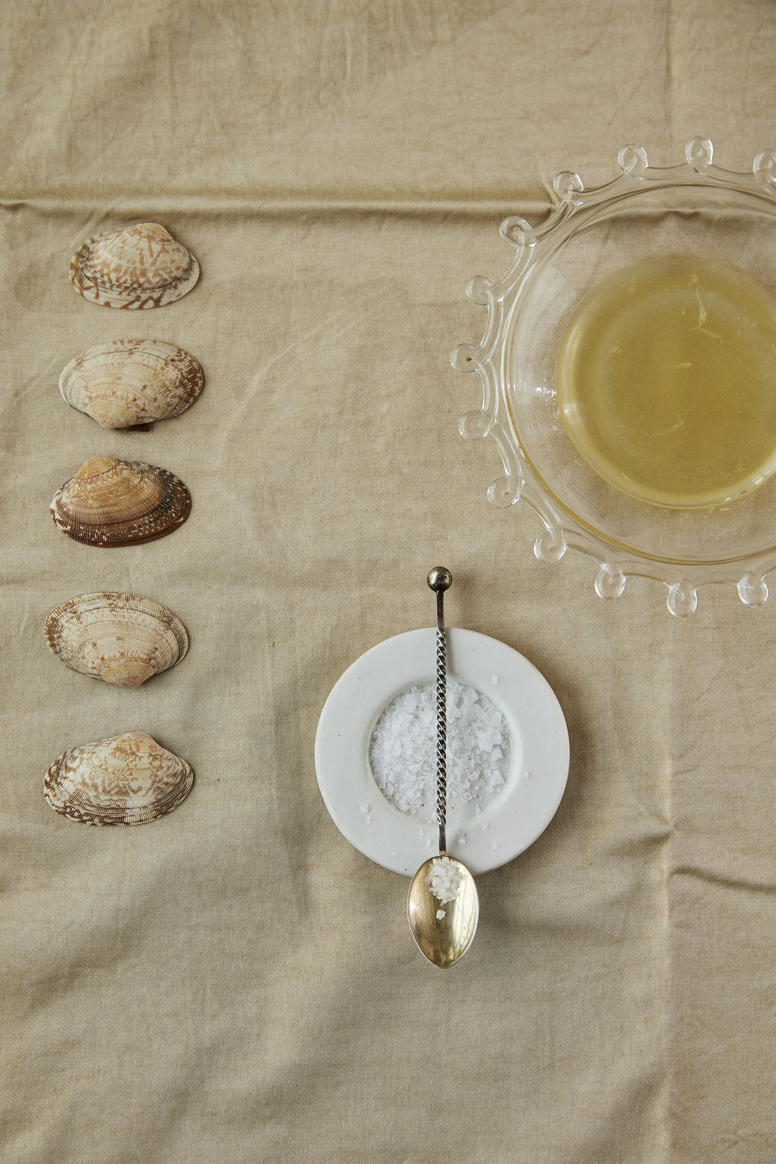 Four seashells lined up on a beige tablecloth, a glass bowl with a yellow-green liquid and a decorative edge, and a small dish with coarse salt and a spoon.