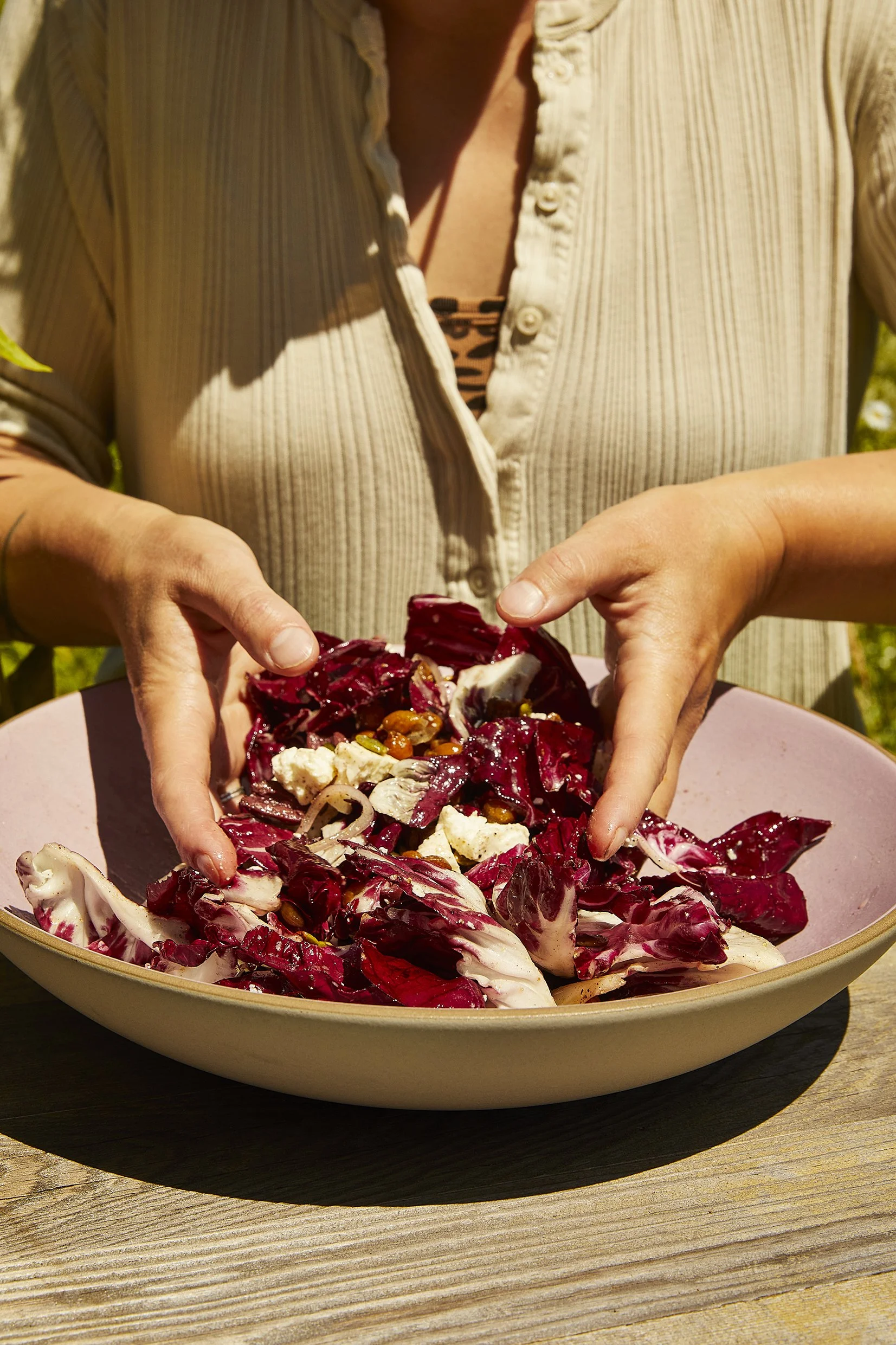 Person in a beige button-up shirt preparing a radicchio salad outdoors on a wooden table.