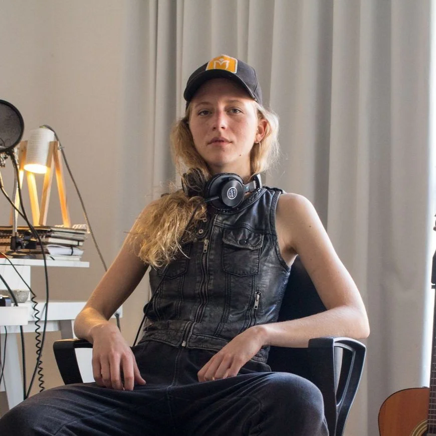 A young woman with blonde hair seated in a black chair in a room with curtains. She is wearing a black leather vest, a black cap with an orange logo, and has a pair of headphones around her neck. There are music equipment and a guitar in the background.