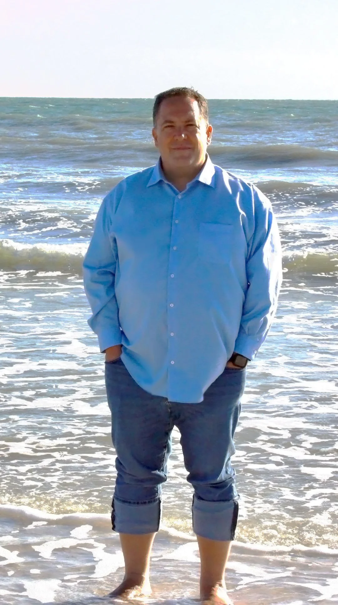 A man in a blue dress shirt and rolled-up jeans standing in the ocean with waves behind him.
