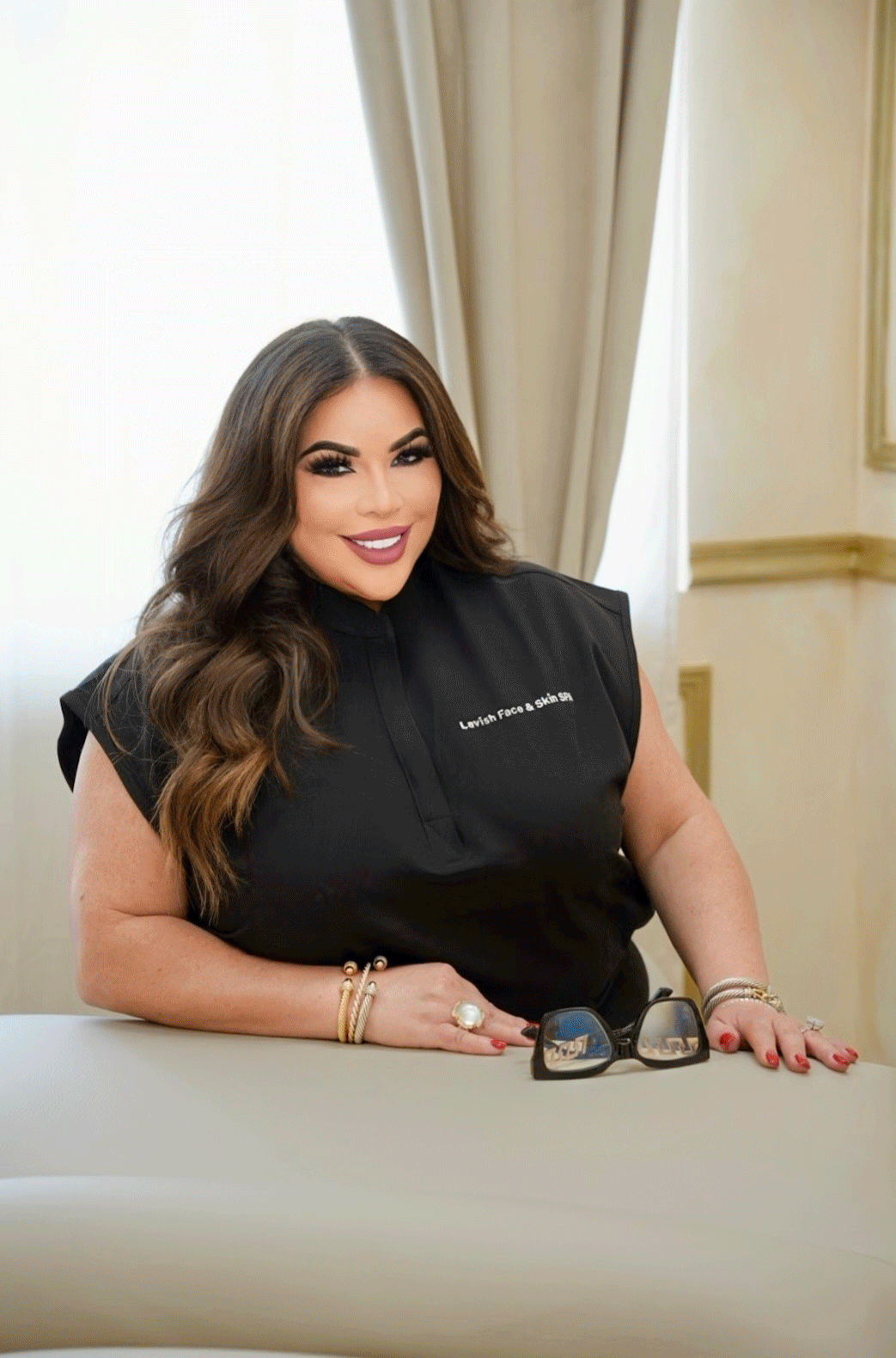 A woman with long brunette hair smiling, sitting at a table with sunglasses in front of her. She is wearing a black top that says "Lavish Face & Skin Spa" on it, and has gold and pearl bracelets and rings on her hands. The background shows beige curtains and a light-colored wall.