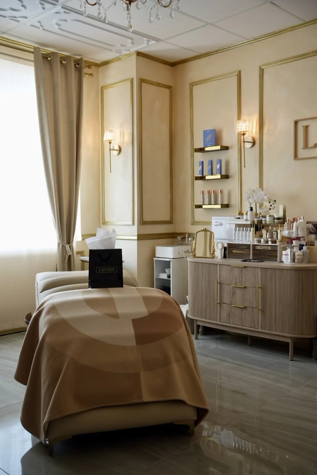 Beauty treatment room with a bed covered in a beige blanket, a black Lavish shopping bag on top, a side cabinet with skincare products, and cream-colored walls with gold trim, sconces, and a chandelier.