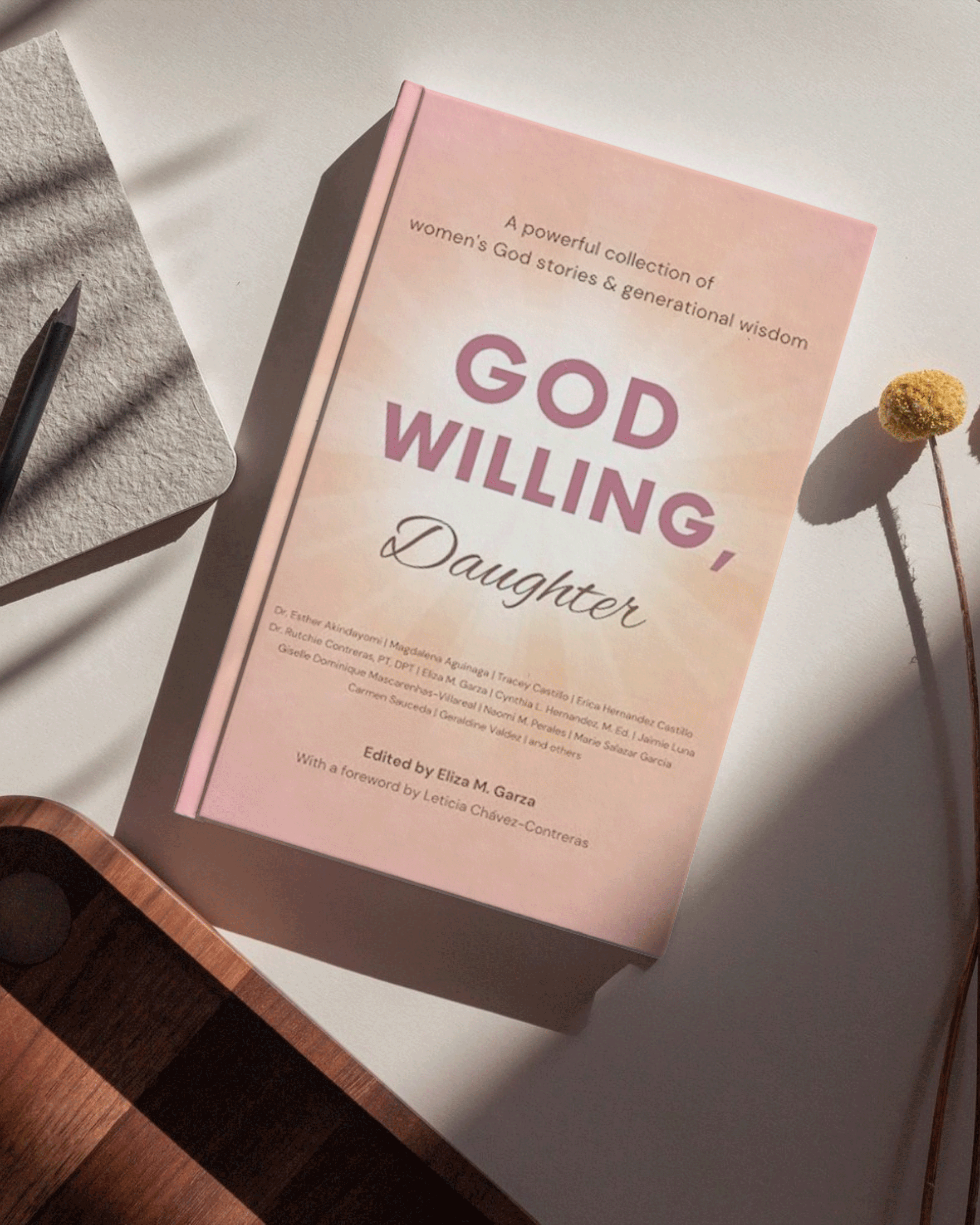 A soft pink book titled 'God Willing, Daughter' is placed on a white surface, next to a dried flower and a gray textured notebook with a pen. The book features a subtitle about women's stories and wisdom.