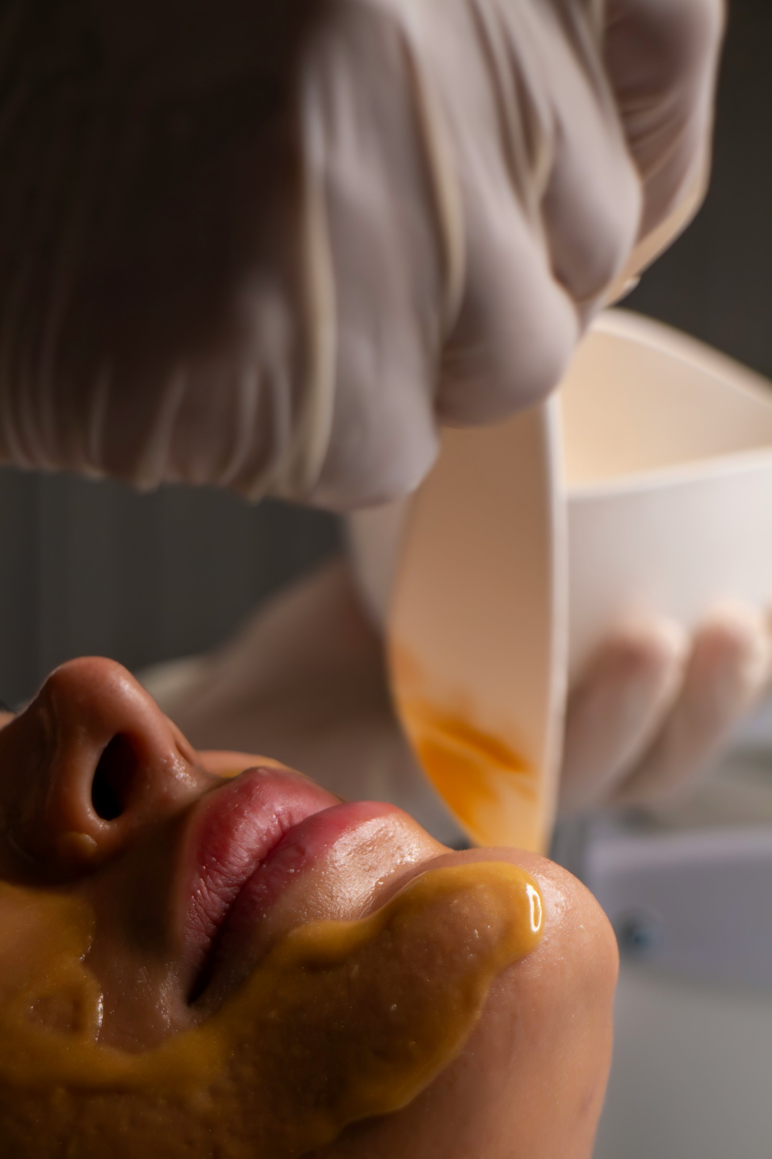 A person with a yellowish substance on their face, lying down with their eyes closed, while another person pours a liquid from a cup near their face.