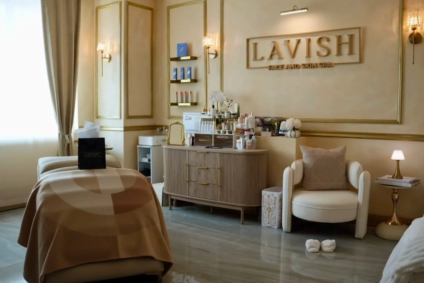 A spa treatment room with a massage bed covered in a towel, a side table with a lamp, a white armchair with a cushion, a cabinet with beauty products, wall-mounted shelves with skincare items, and a large sign saying "LAVISH FACE AND SKIN SPA" on the wall.