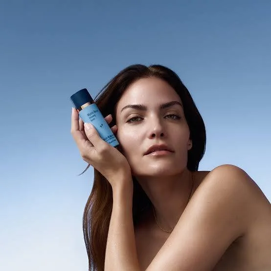 A woman with long brown hair holding a blue skincare product bottle against her temple, against a blue sky background.