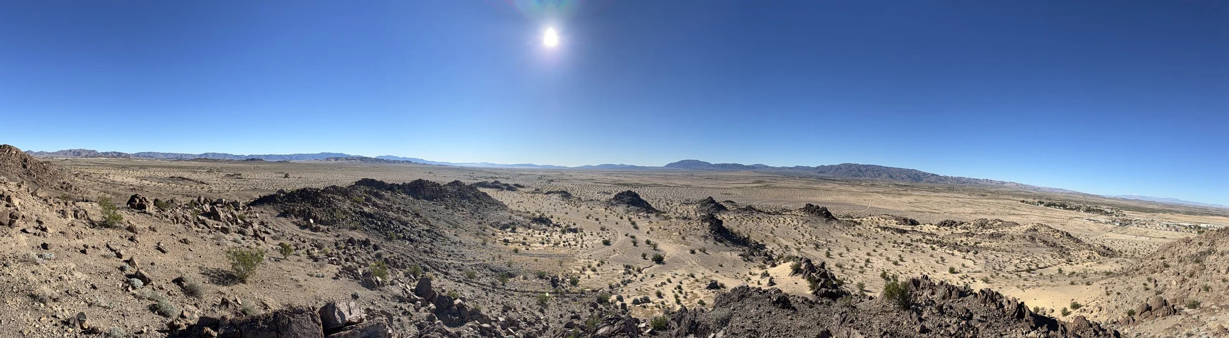 Panoramic view of a desert landscape with rocky hills, sparse vegetation, and distant mountains under a bright blue sky with the sun