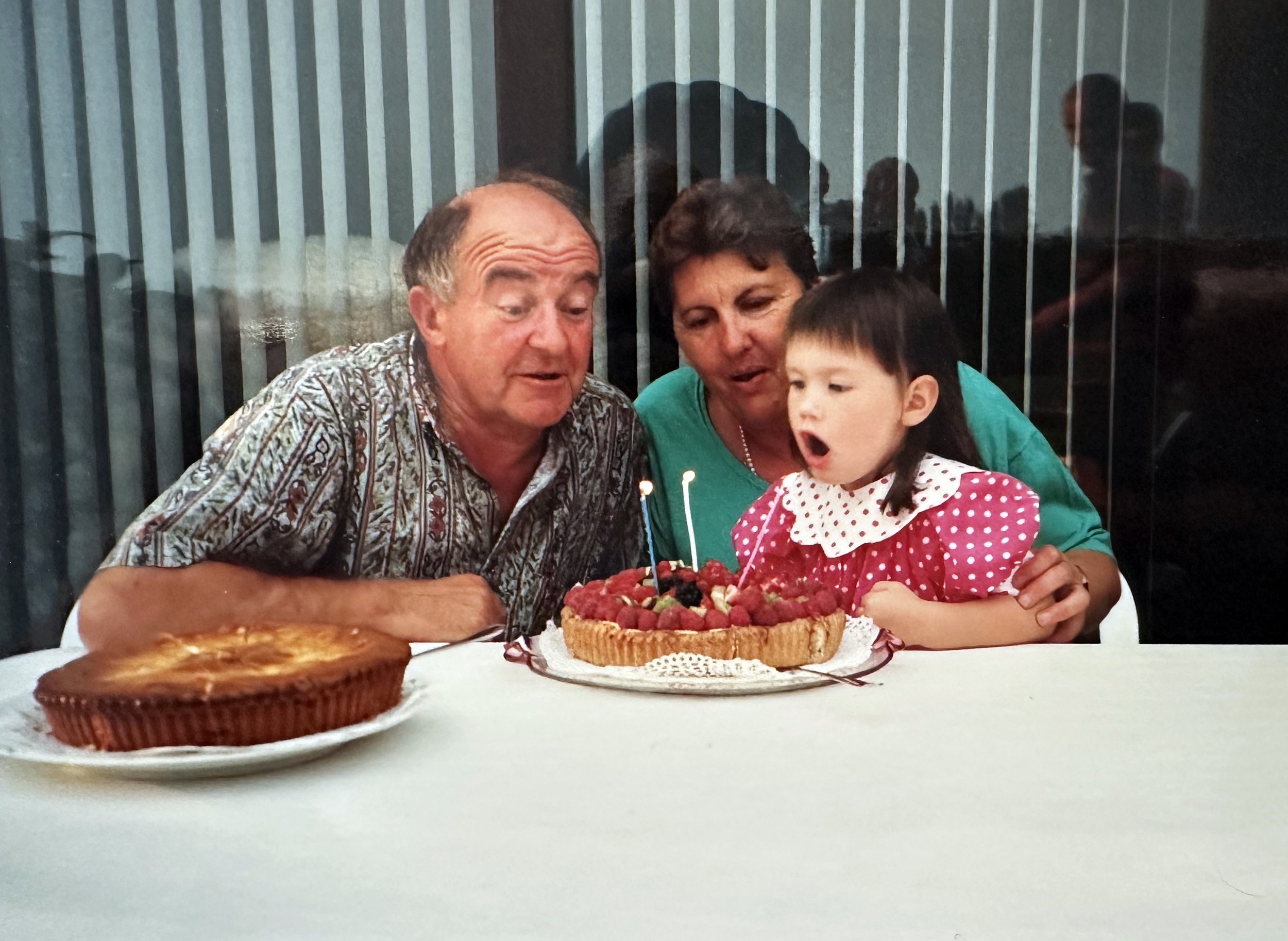 Een familie viert een verjaardag met een taart met kaarsjes, omringd door drie volwassenen en een meisje in een roze jurk met witte stippen.