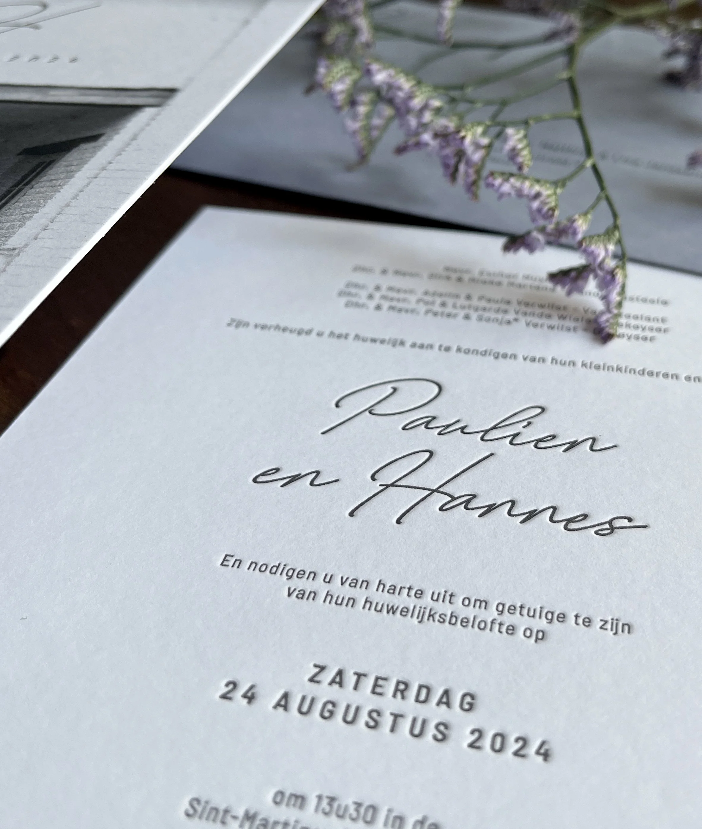 Een huwelijksuitnodiging op een tafel met een paar paarse bloemen eroverheen gelegd.