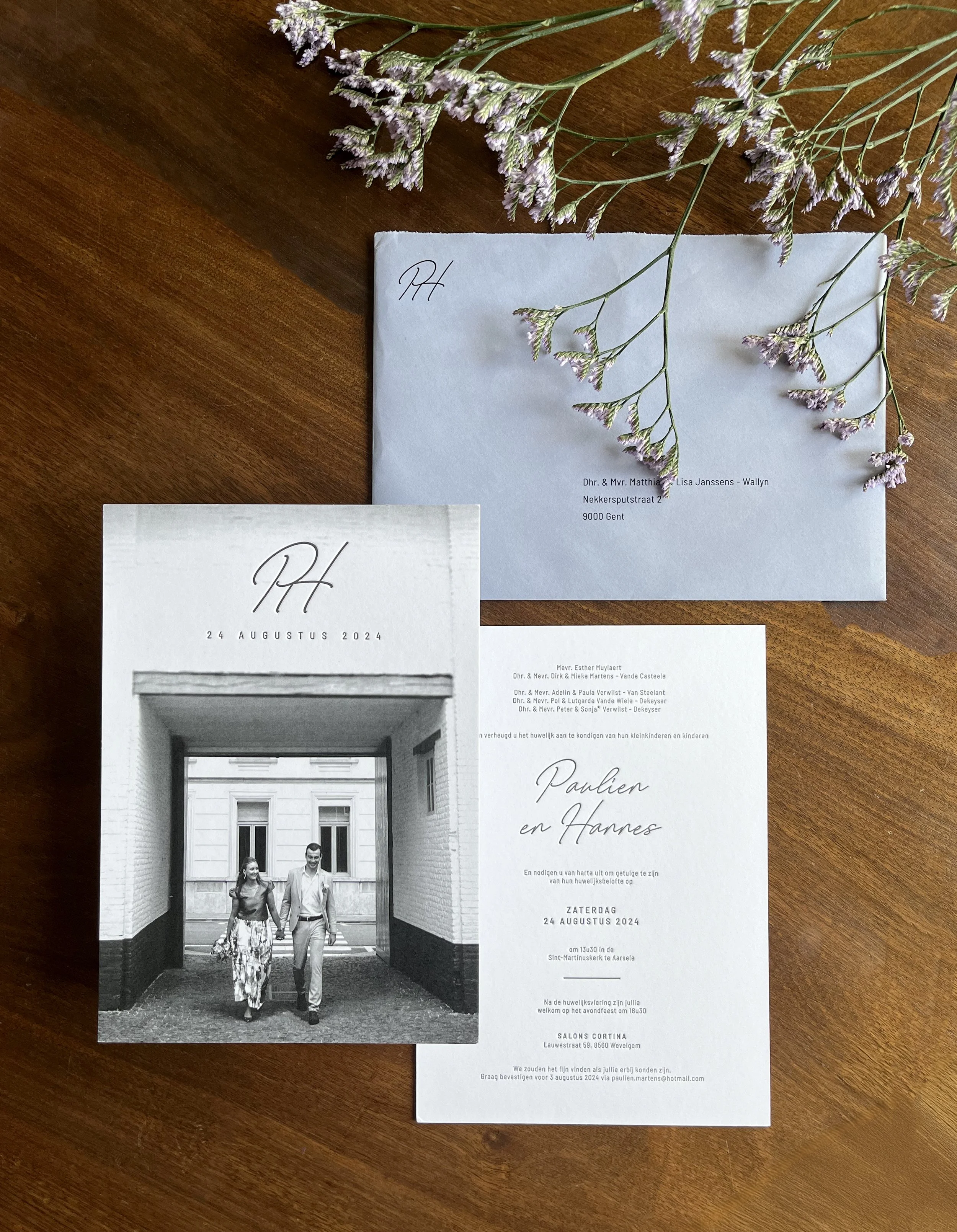 Een uitnodiging voor een bruiloft met een zwart-wit foto van een paar dat door een poortje loopt, bloemen en een envelop op een houten tafel.