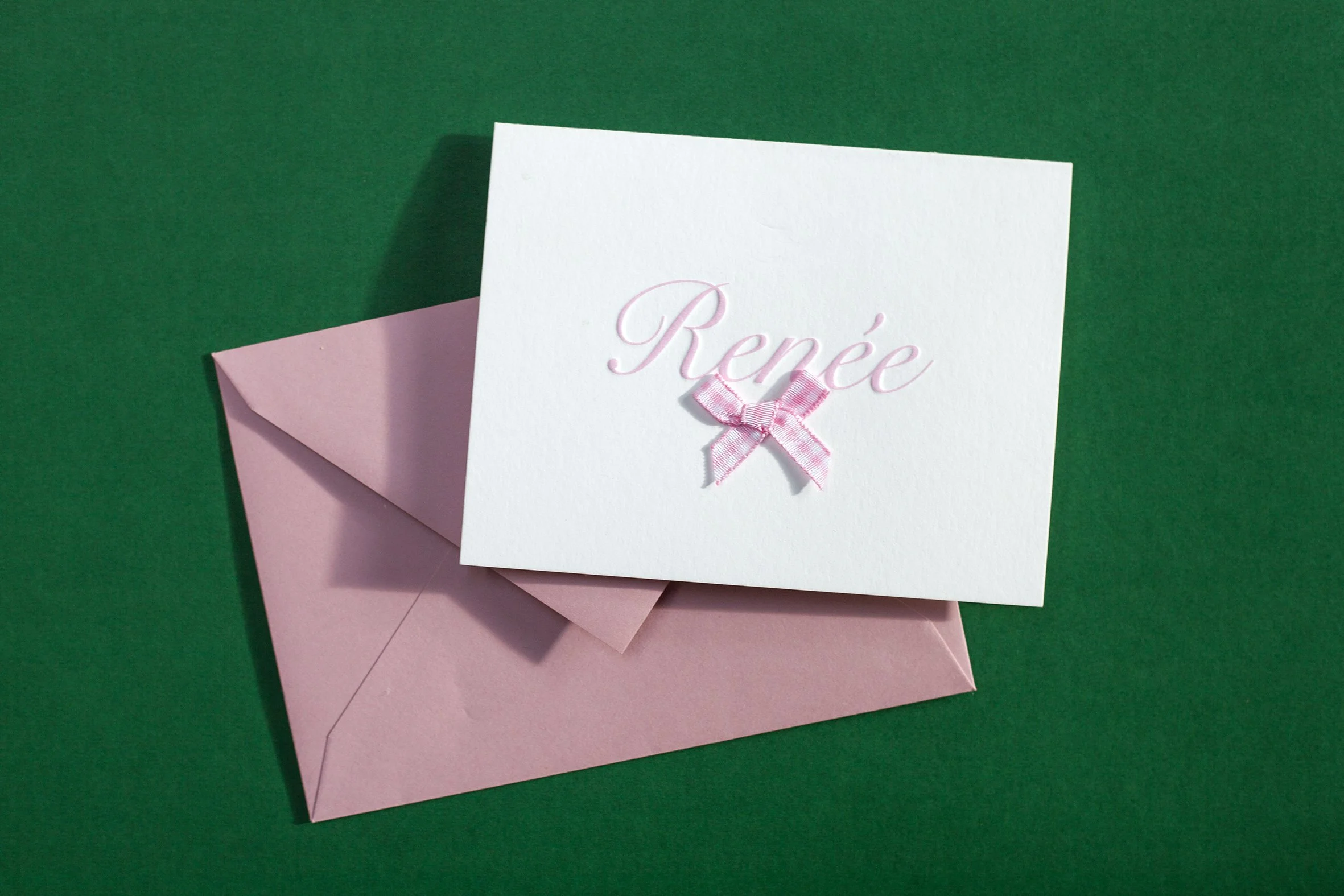 Witte kaart met de naam Renée in letterpress en een roze strik, op een roze envelop op een groene achtergrond.