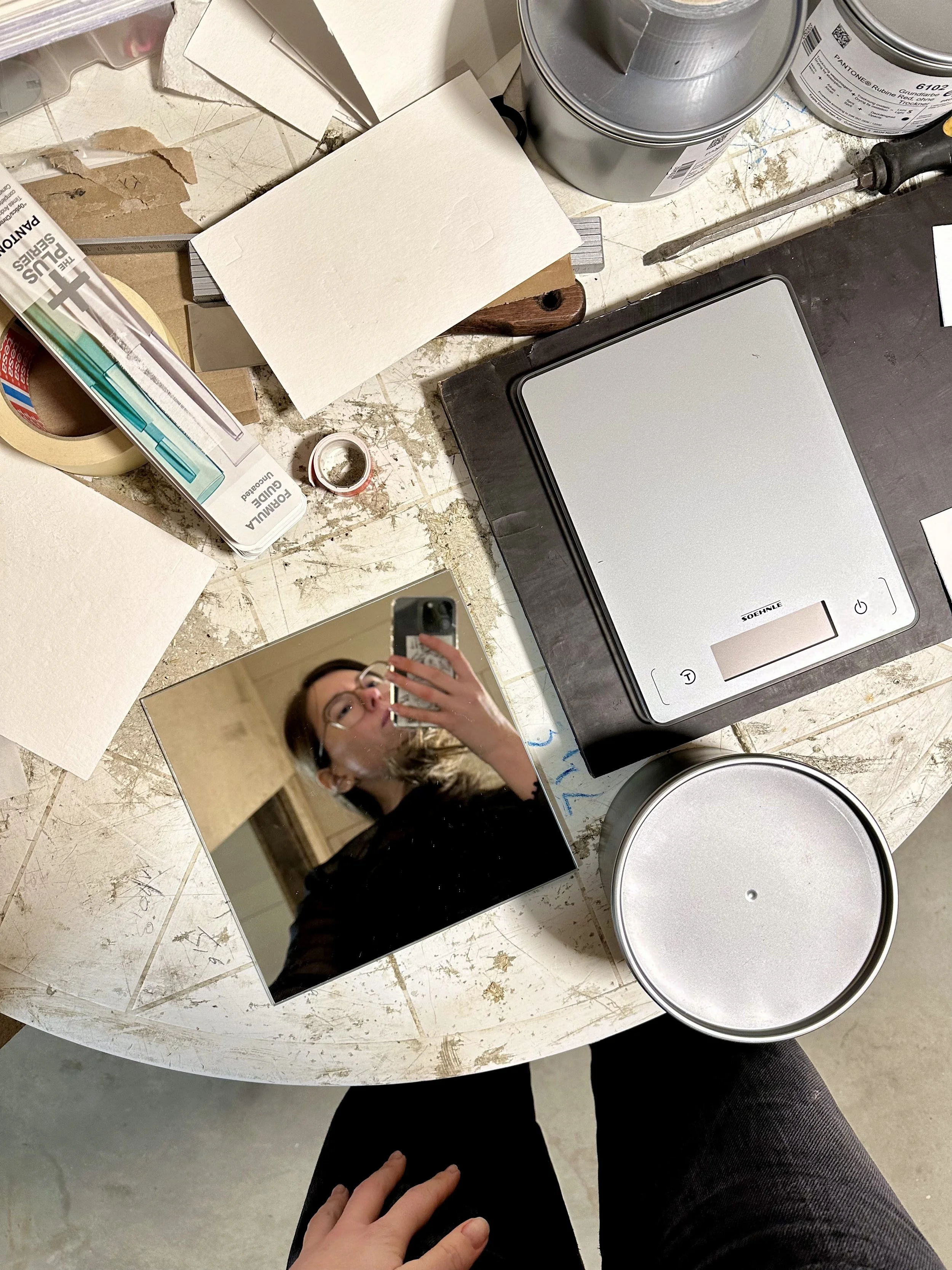 Een collage van een tafel met verschillende materialen zoals tape, verfpotten, een digitale weegschaal en een spiegel met een vrouw die selfie neemt.