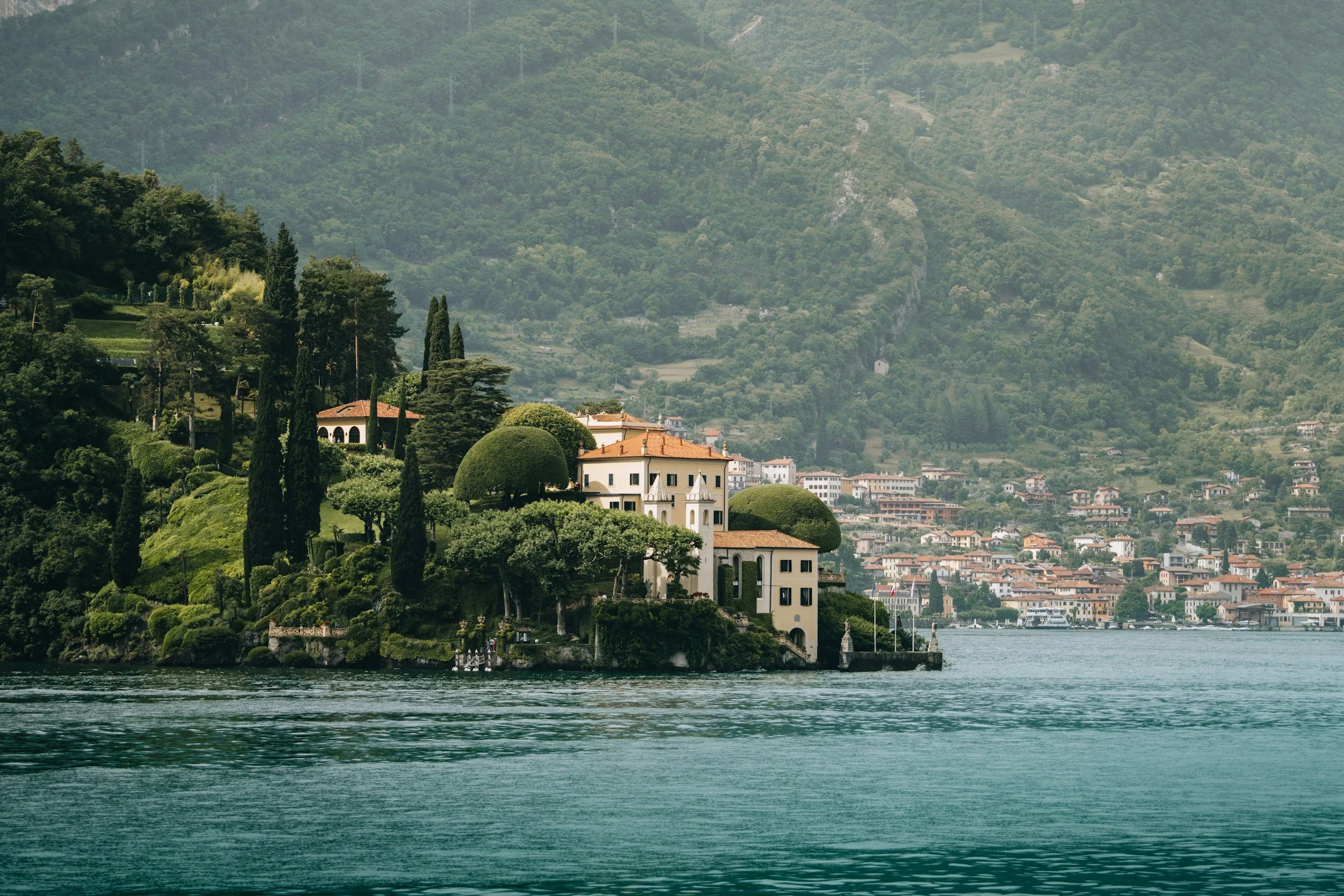 A scenic lakeside view featuring a large house with a red-tiled roof surrounded by lush green trees and shrubbery, with a hillside village in the background and calm water in the foreground.