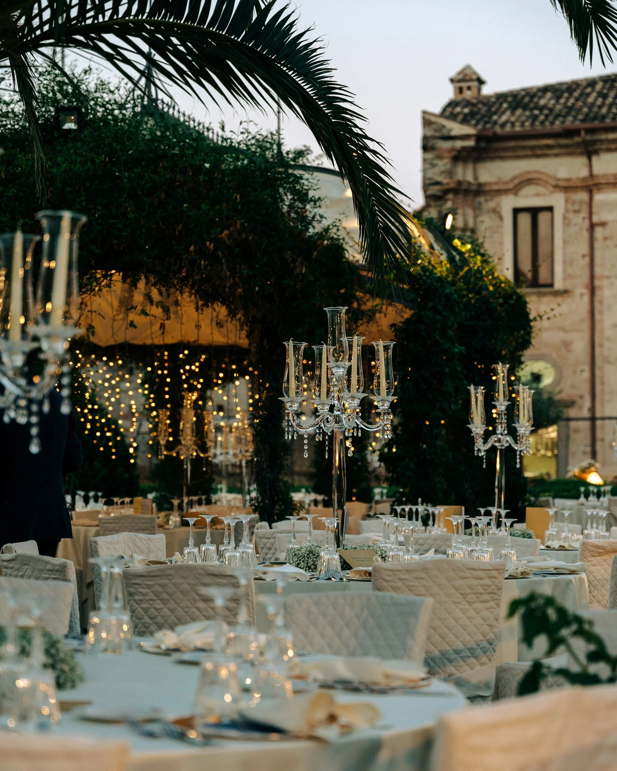 Elegant outdoor wedding reception with round tables, white tablecloths, and tall chandeliers, surrounded by lush greenery and an historic stone building at dusk.