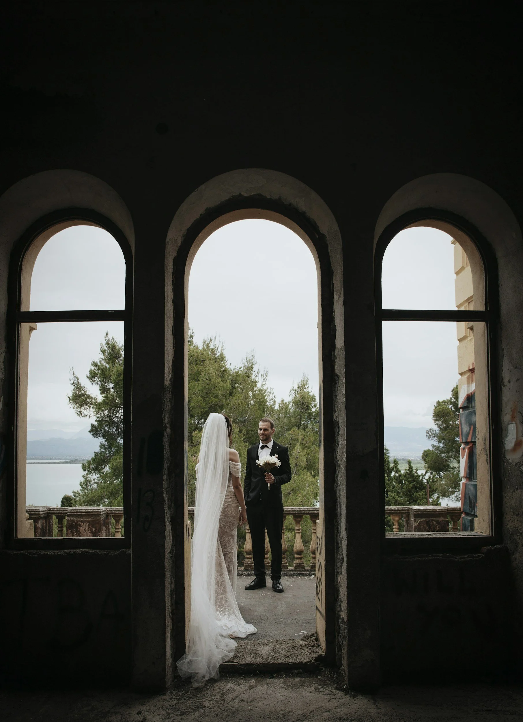 A bride and groom in wedding attire standing outdoors, framed by three arched windows, with trees and a body of water in the background.