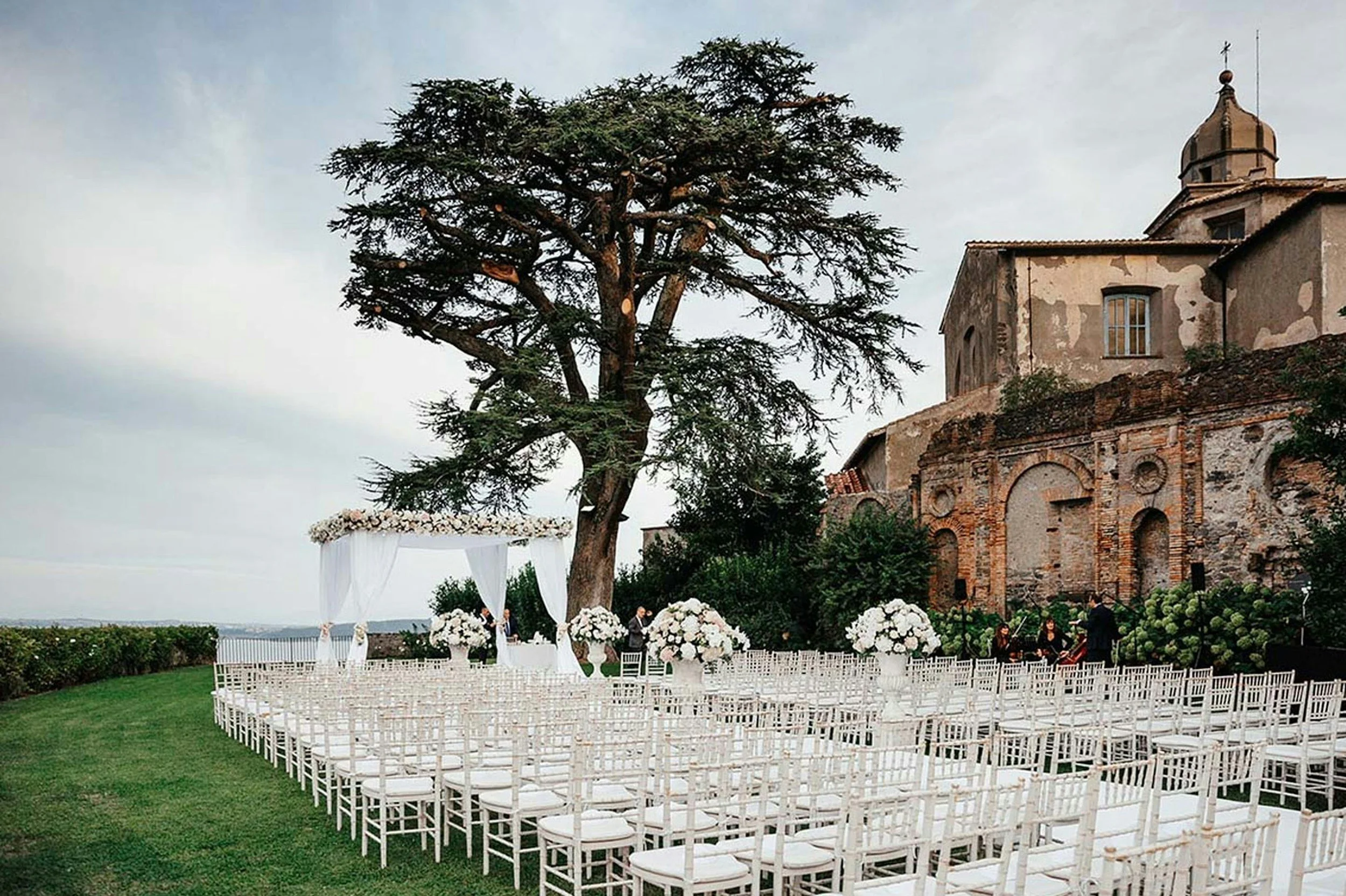 Outdoor wedding setup with white chairs arranged in rows facing a decorated altar, surrounded by large floral arrangements, under a big tree with an old stone building in the background.