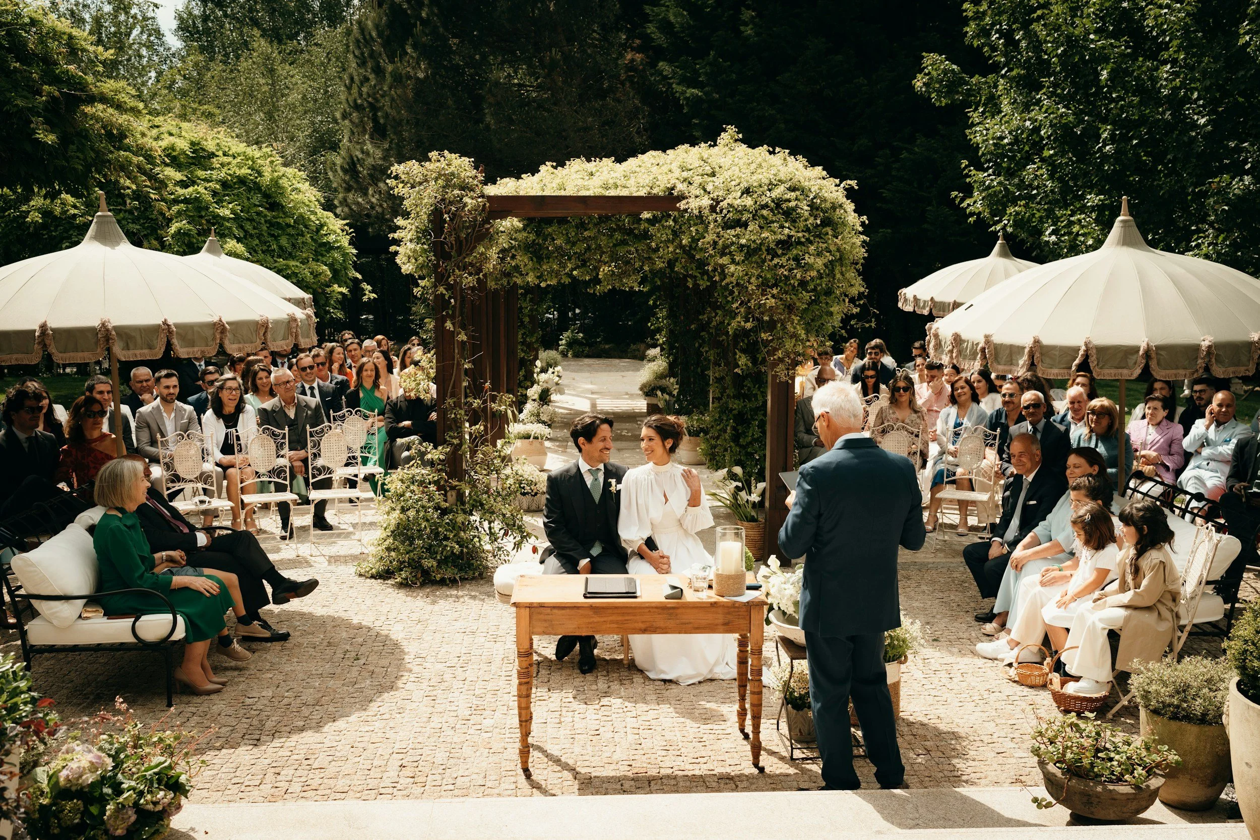 A wedding ceremony outdoors with guests seated under umbrellas, a couple sitting at the altar, and an officiant standing in front of them, in a lush garden setting.